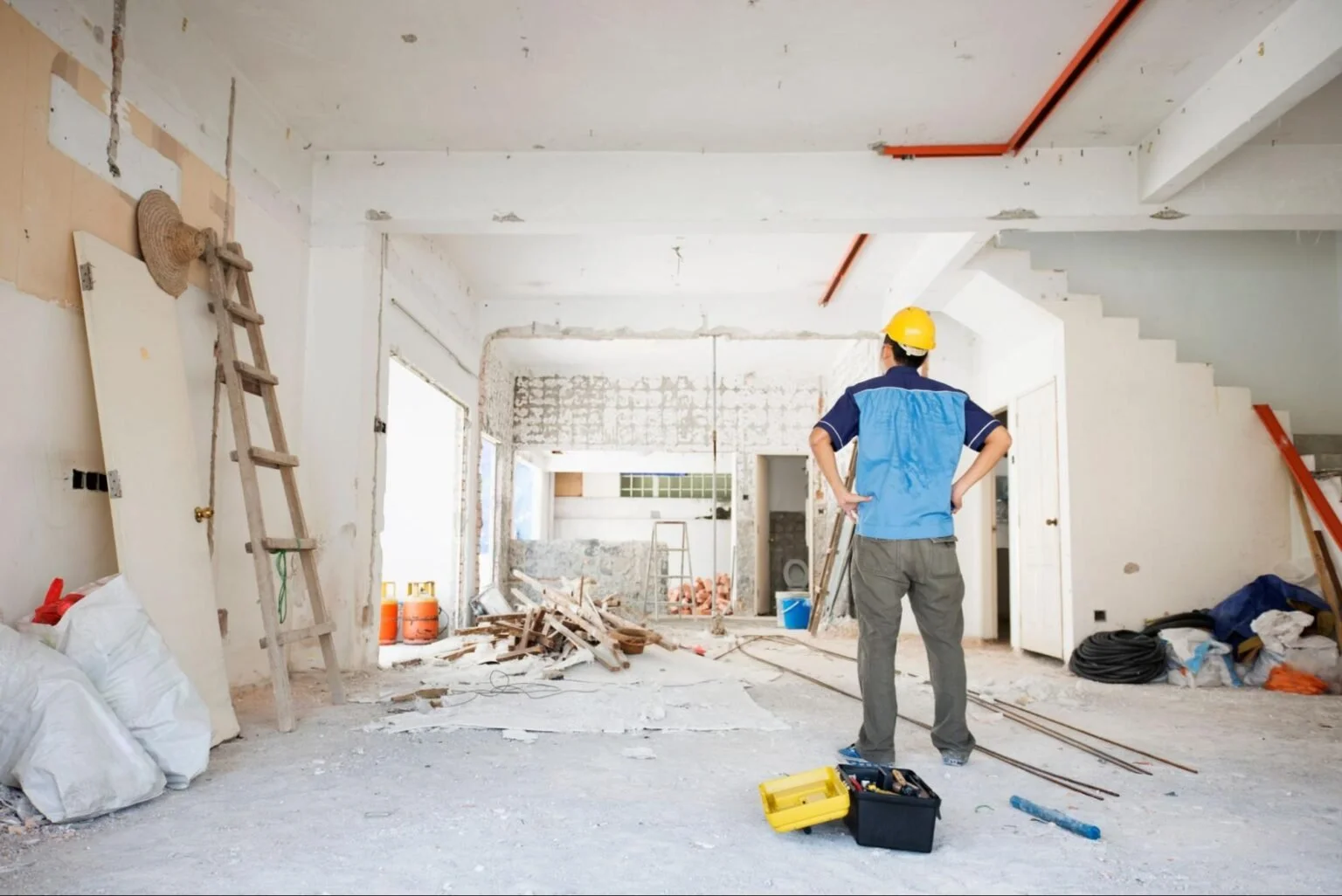 Construction worker wearing a yellow helmet standing in a room under renovation with hands on hips and construction tools on the ground.