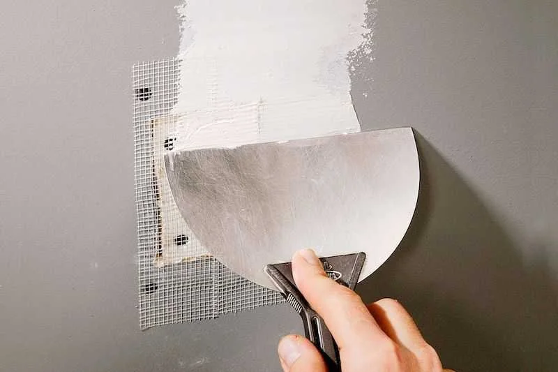 A person using a trowel to apply a patch of drywall compound to a wall with visible mesh tape.