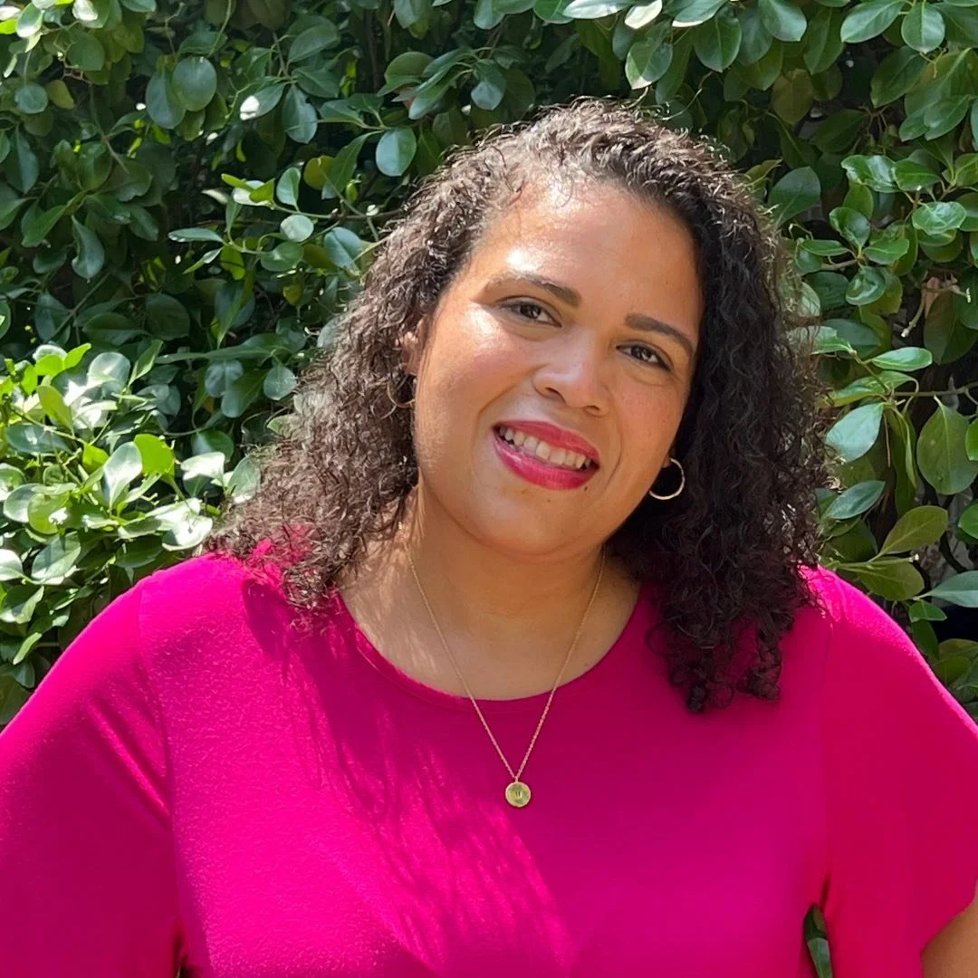 A woman with curly dark hair smiling in front of green leafy bushes, wearing a bright pink top, hoop earrings, and a gold necklace.