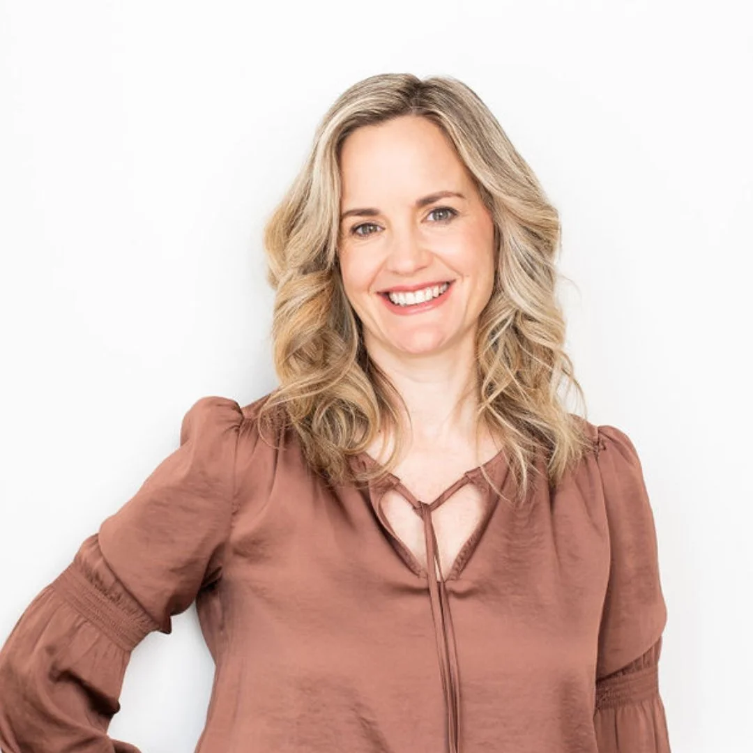 A smiling woman with blonde, wavy hair wearing a brown blouse, standing against a white background.