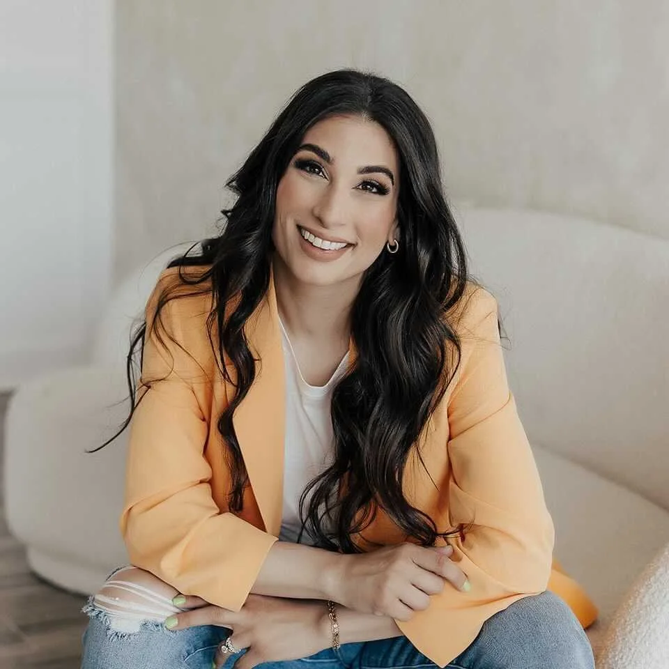 A woman smiling while sitting on a couch, wearing a yellow blazer over a white top, with long black hair and makeup.