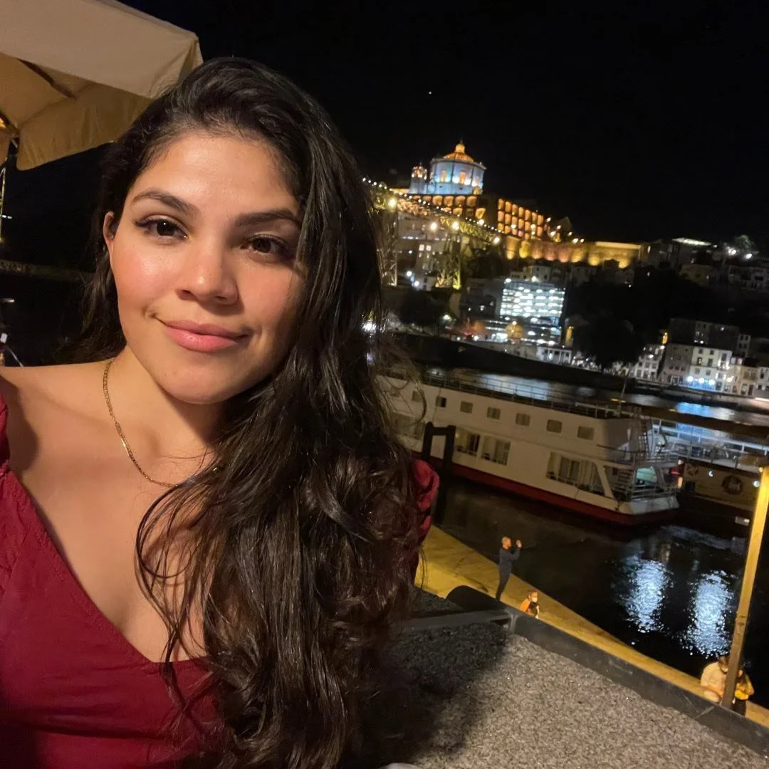 A young woman with long dark hair smiling at night with a cityscape and docked boat behind her.