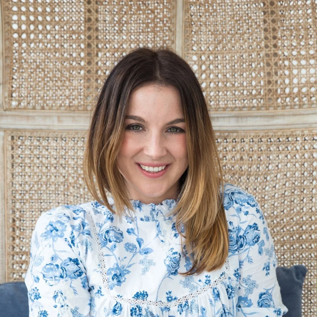 A young woman with shoulder-length brown hair and blue eyes, smiling, wearing a white blouse with blue floral patterns, sitting in front of a woven rattan backdrop.