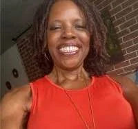 Smiling woman taking a selfie, wearing a red sleeveless top, with a brick wall and outdoor setting in the background.