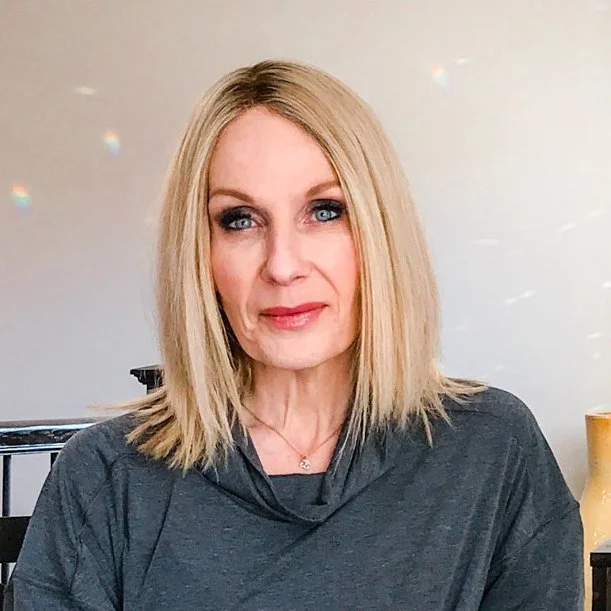 A woman with blonde hair and blue eyes wearing a dark gray top, sitting indoors against a plain light-colored wall.
