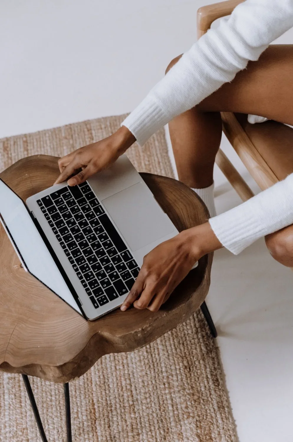 Person in white sweater typing on a laptop placed on a wooden side table.