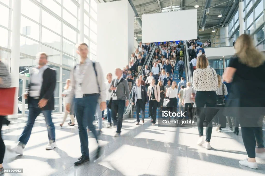 Crowd of people walking through a modern, glass-enclosed indoor space with stairs and an escalator.