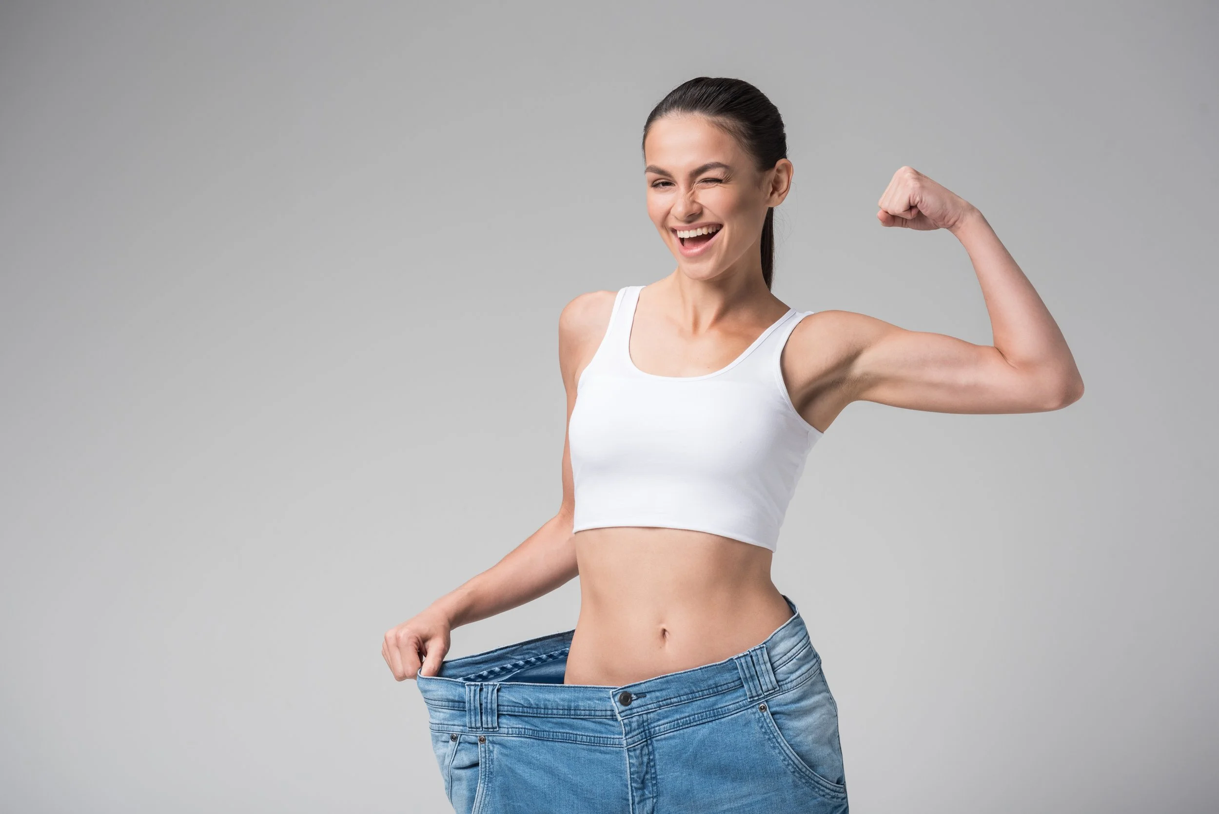 A woman smiling and wearing a white crop top and large jeans, indicating she has lost weight.