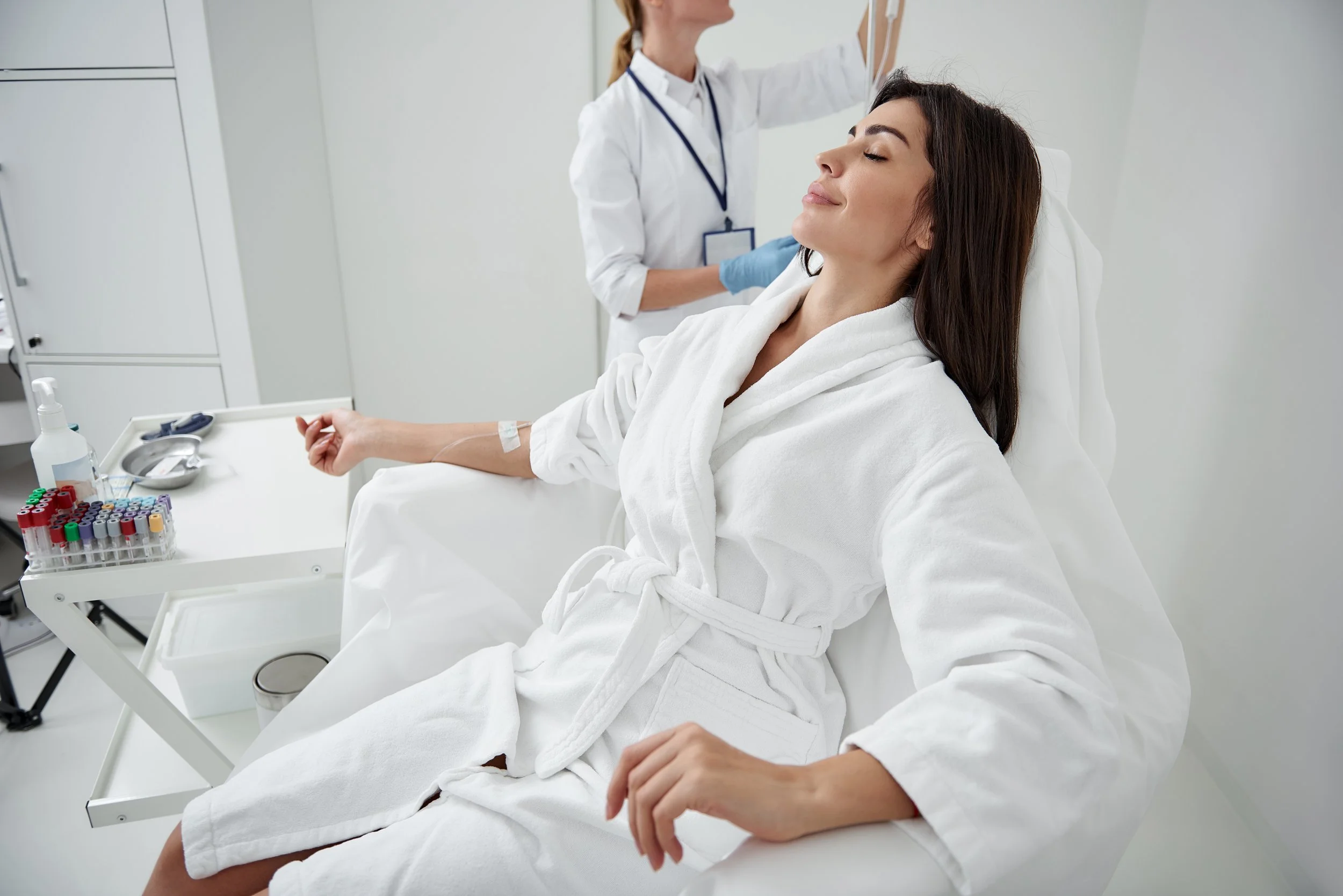 A woman in a white bathrobe undergoing a medical procedure, sitting on a medical examination chair with her arm extended. A nurse or doctor stands behind her in a clinical setting, preparing or administering a treatment.