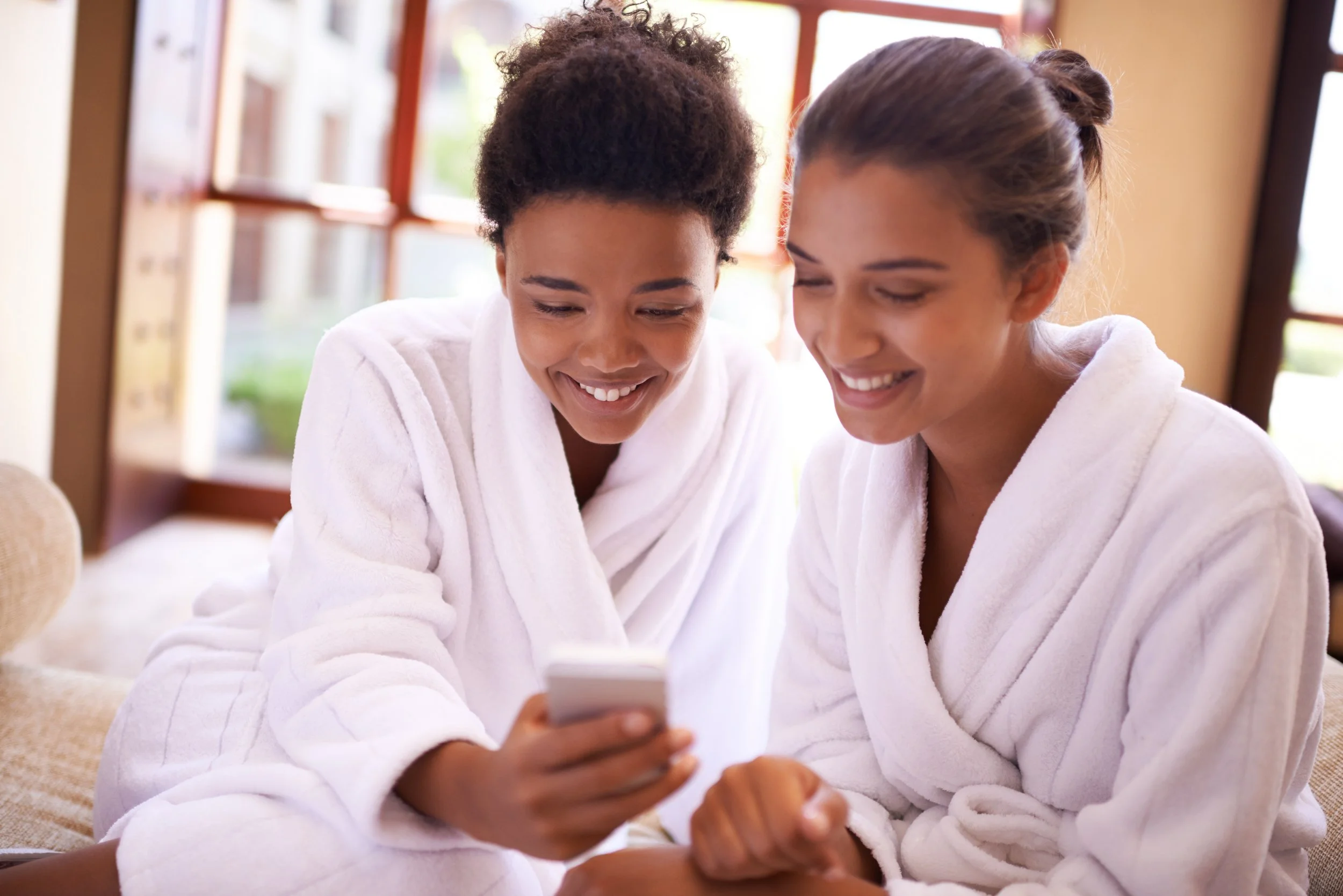 Two women in white robes smiling and looking at a smartphone together indoors near a window.