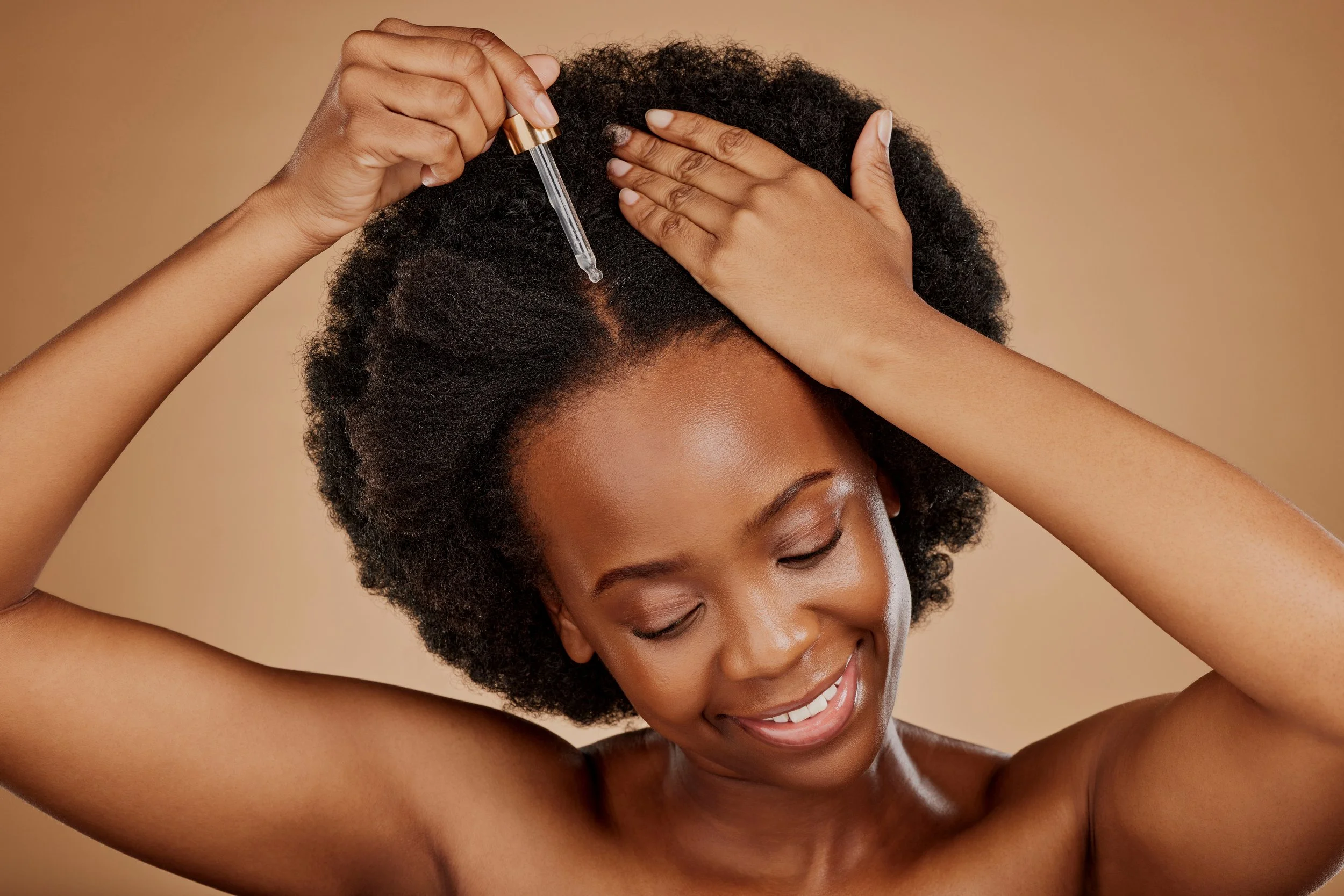 A woman with natural curly hair smiling with her eyes closed, as she applies a dropper of serum to her scalp, holding the dropper in her right hand and using her left hand to part her hair.