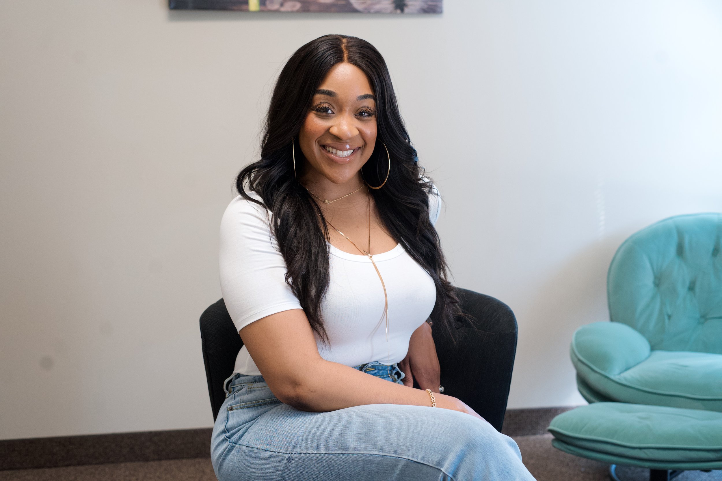 A smiling woman with long black hair, wearing a white t-shirt, light blue jeans, hoop earrings, and layered necklaces, sitting on a black chair in a room with a white wall and teal chair.