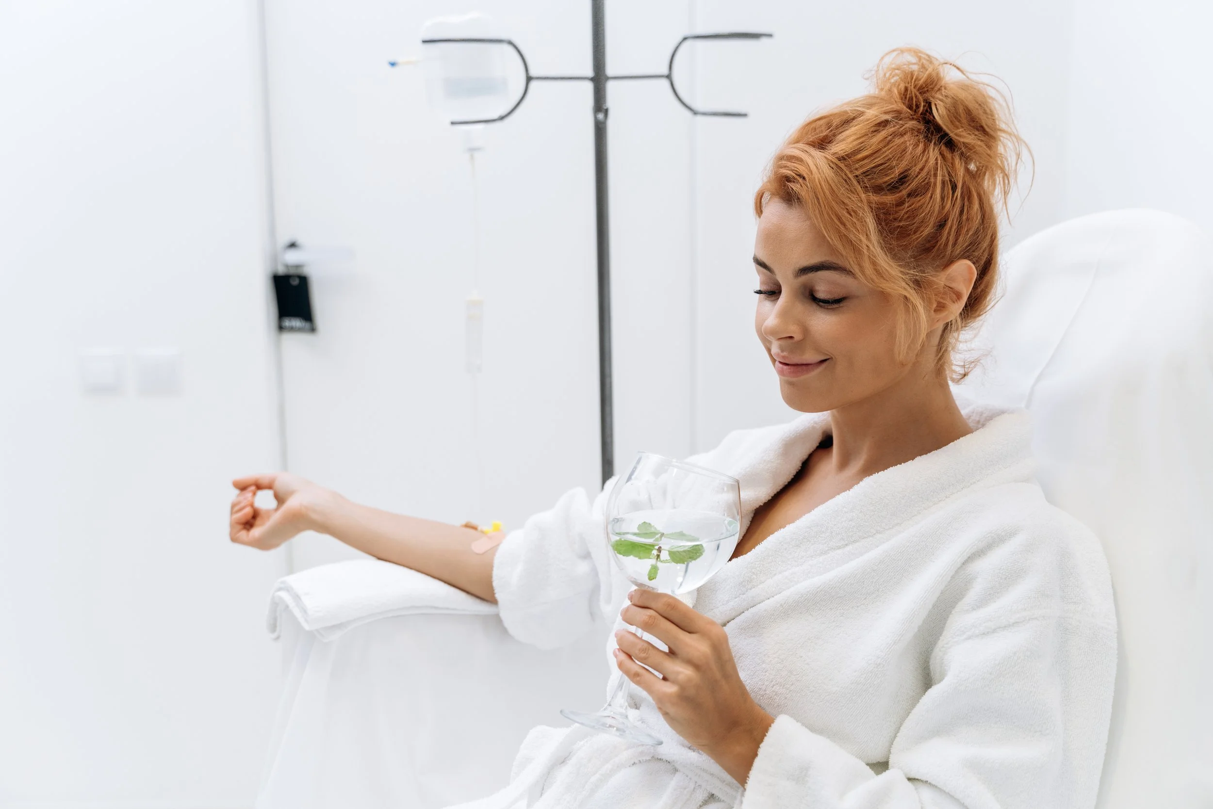 A woman with red hair in a white bathrobe sitting in a hospital bed, holding a glass of water with mint leaves, with an IV in her arm, smiling gently.