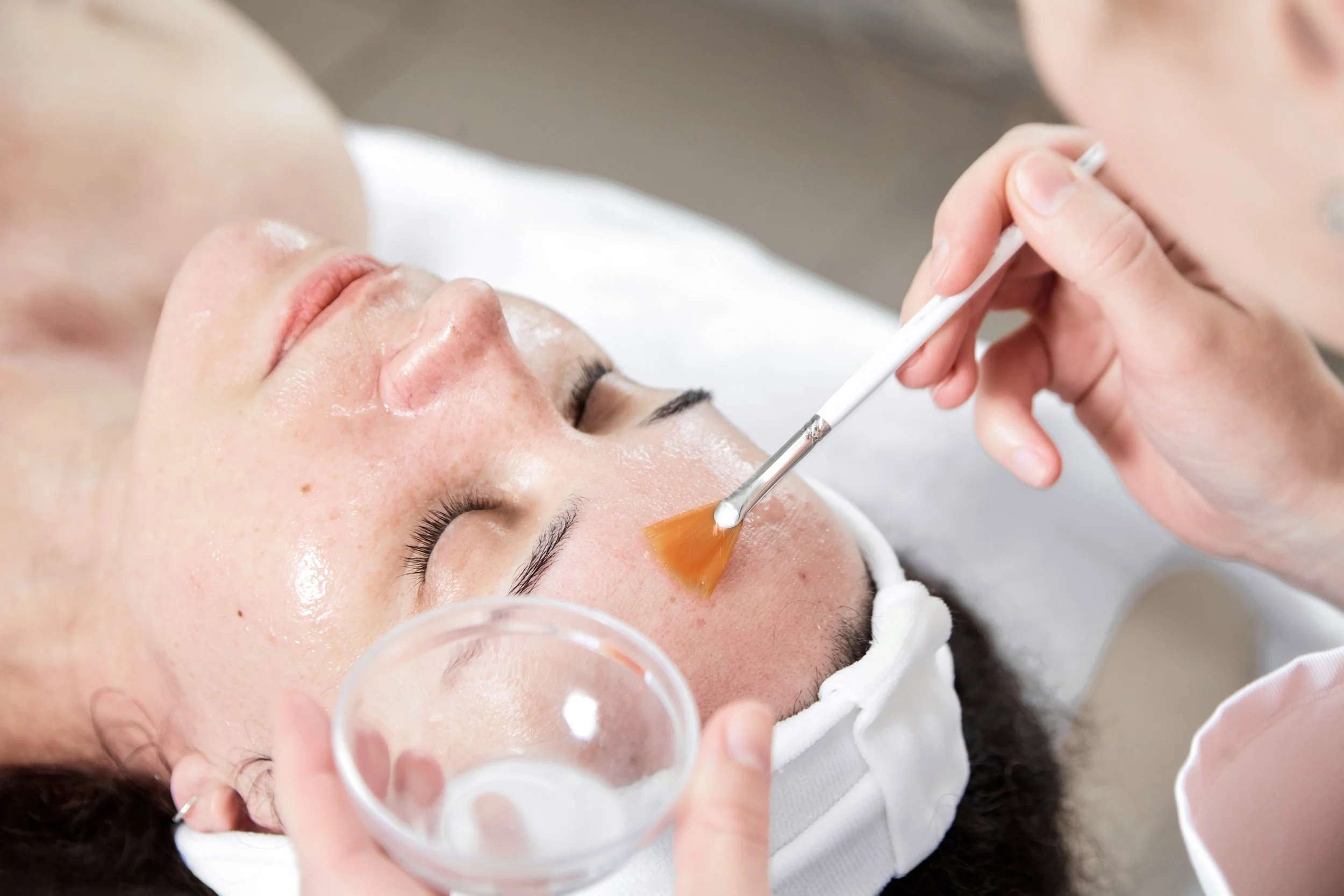 A woman receiving a facial treatment in a spa or skincare clinic, with a skincare professional applying a product to her face using a brush.