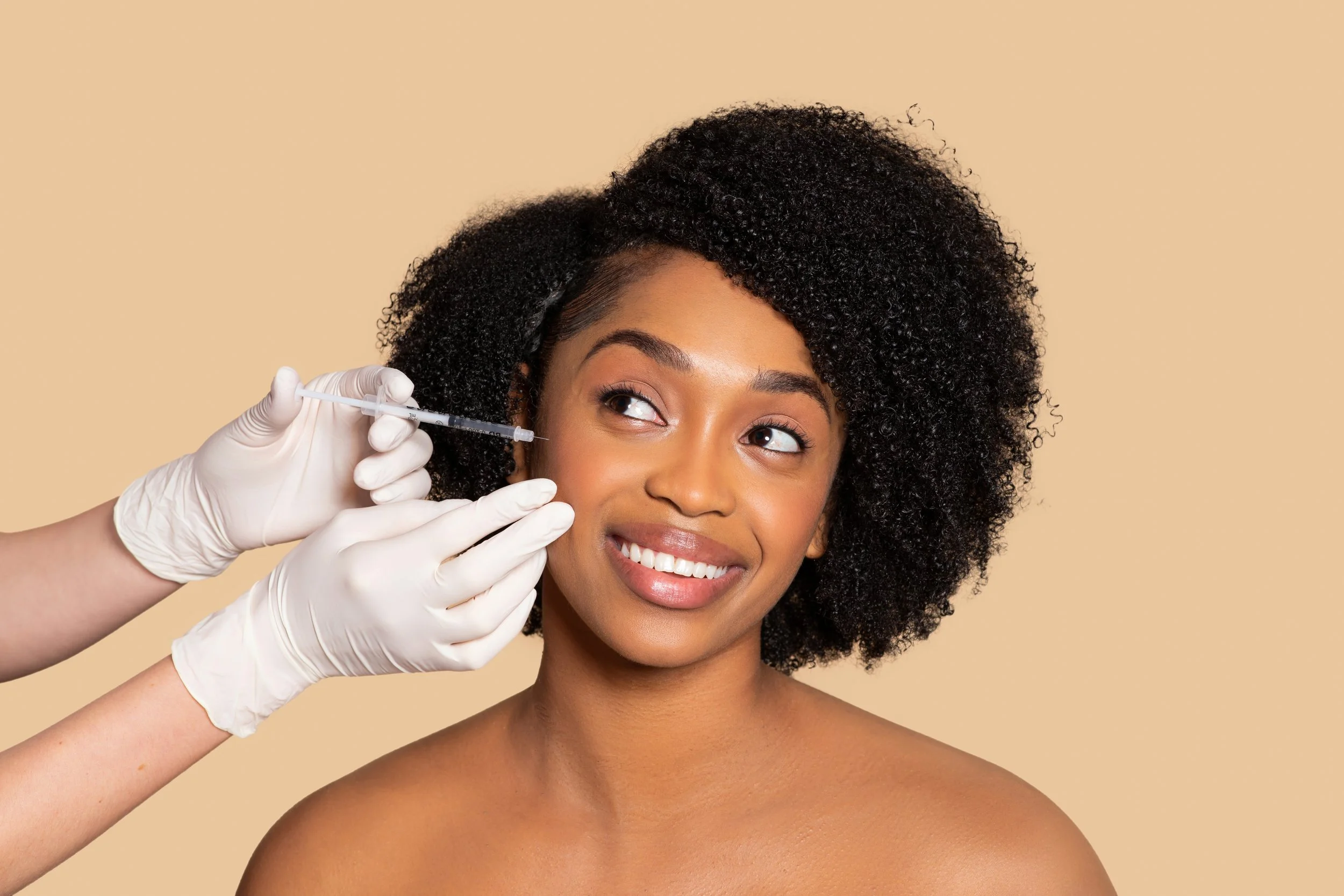 A woman receiving a cosmetic injection in her cheek from a medical professional wearing white gloves, with a neutral beige background.