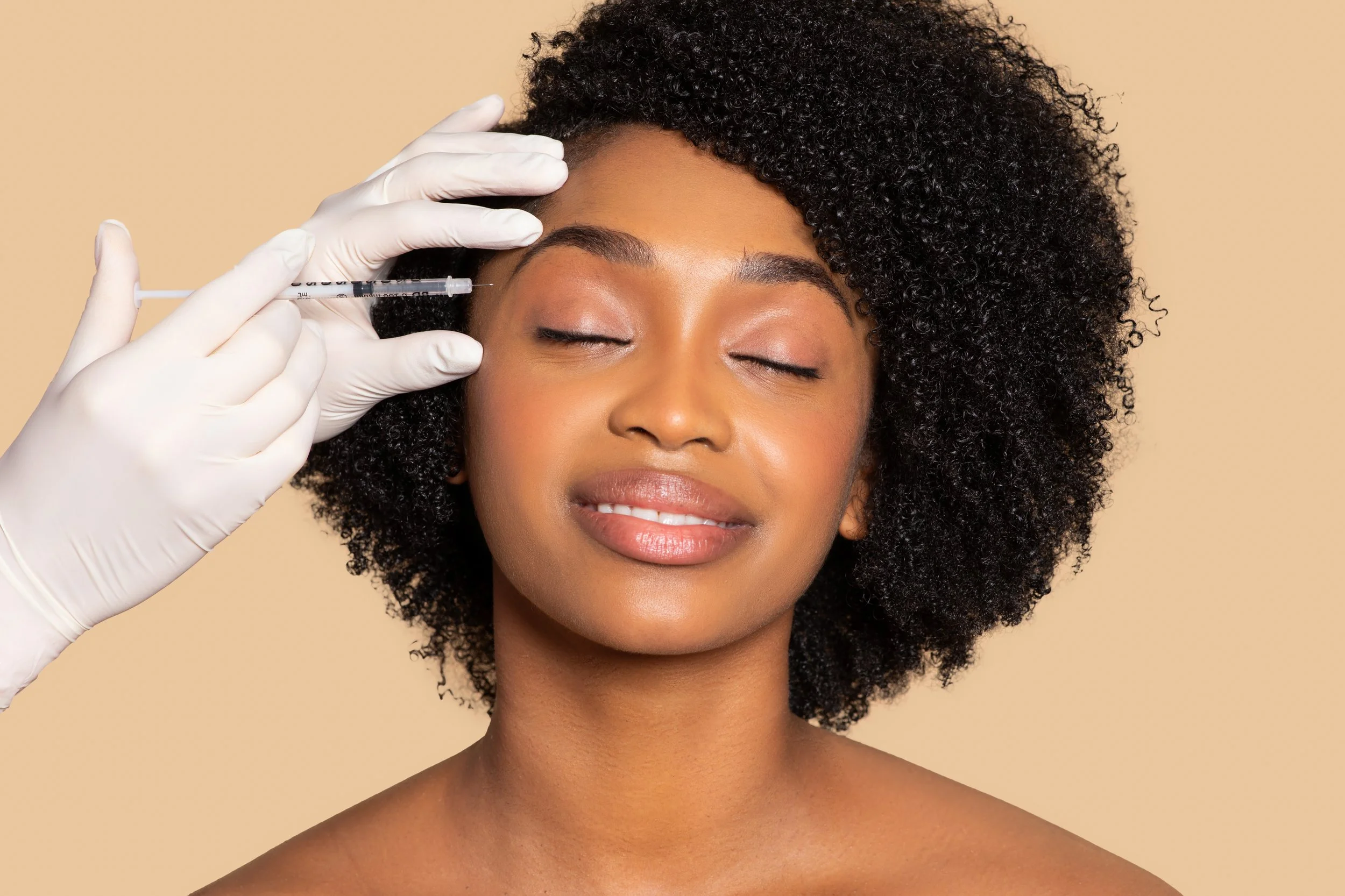 A woman with curly black hair receives an injection in her forehead from a medical professional wearing glove, with her eyes closed and a neutral expression.