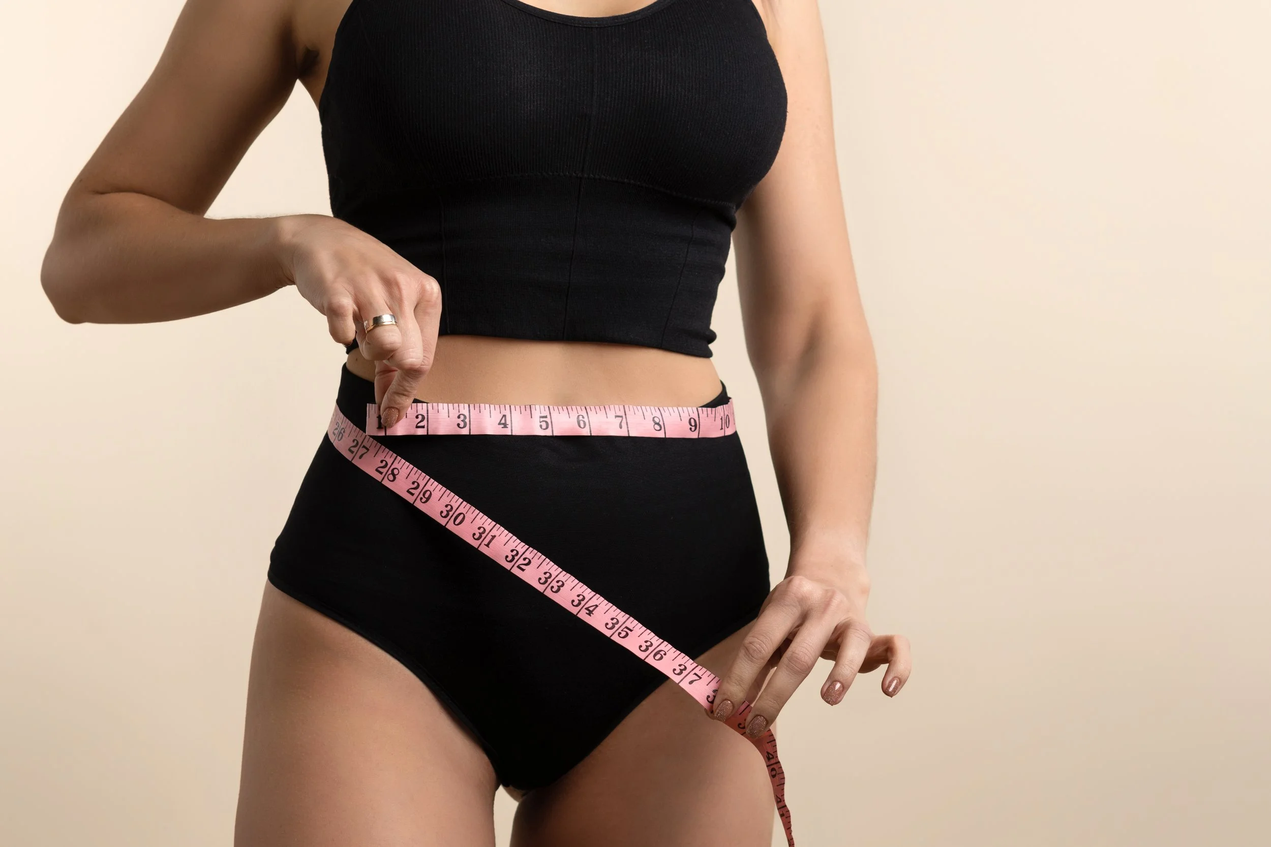 A woman in black workout clothes measuring her waist with a pink measuring tape.