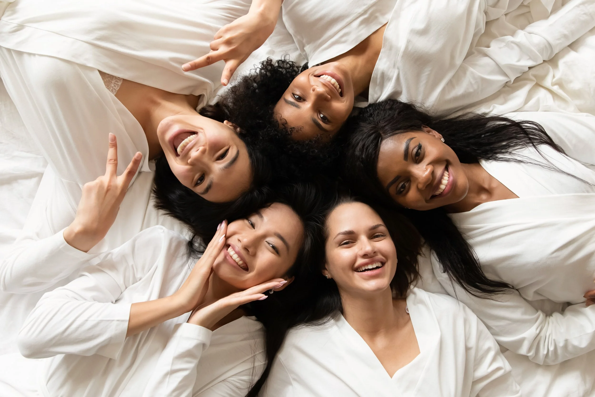 Six women lying in a circle on a bed, smiling and making peace signs.