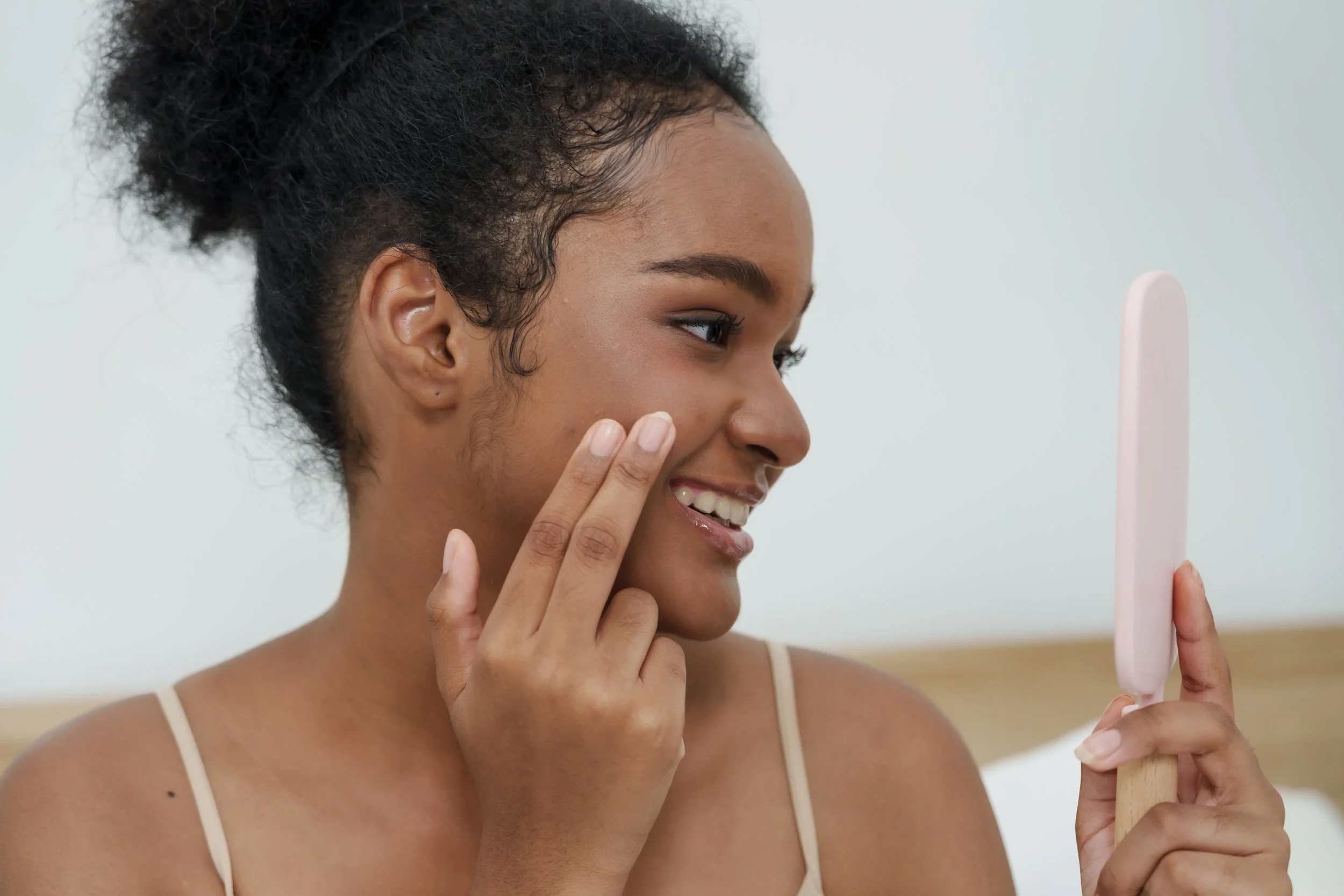 A woman smiling while looking at herself in a mirror, gently touching her cheek with her hand.