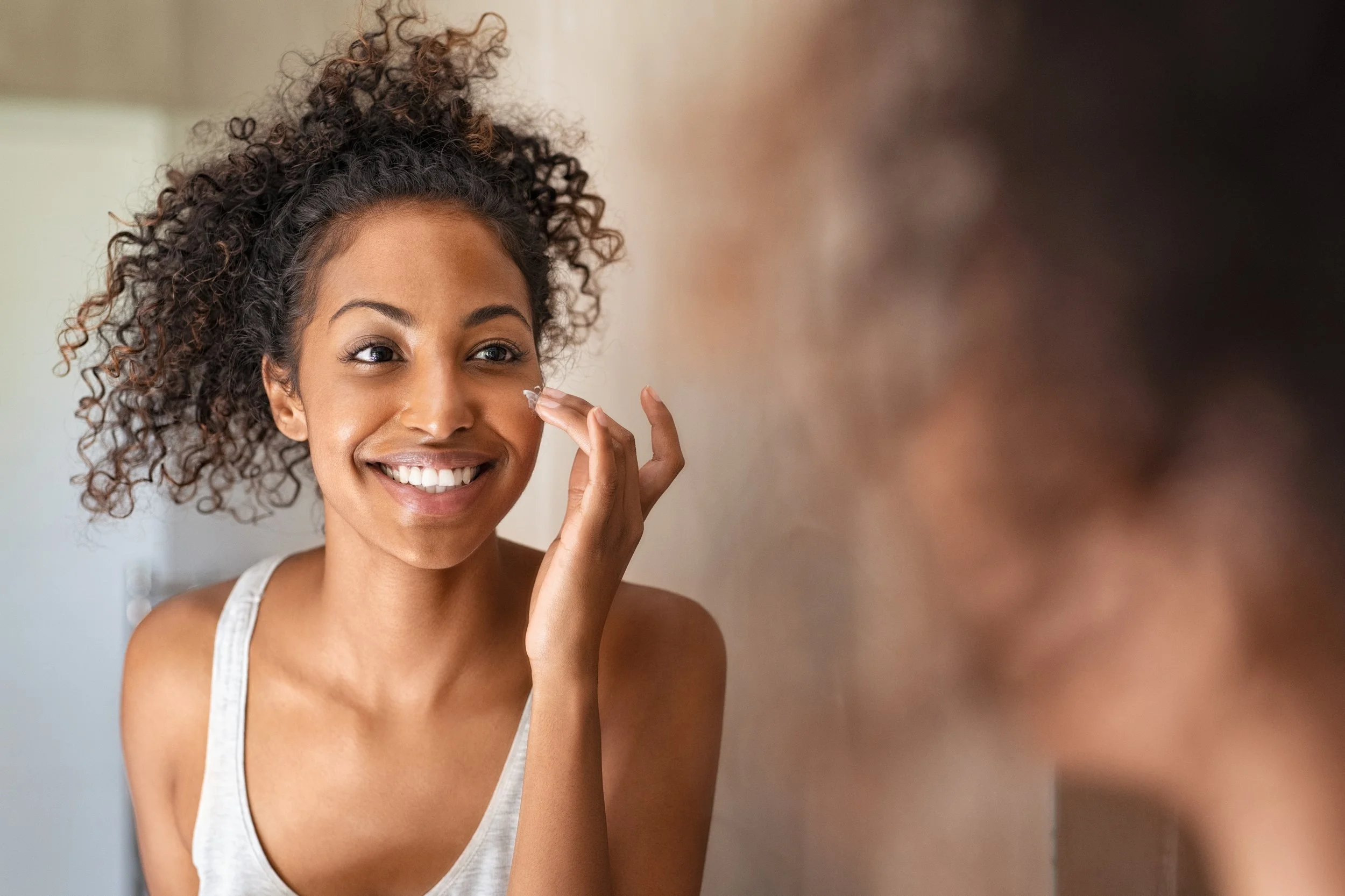 A woman with curly hair smiling and touching her face while looking at her reflection.