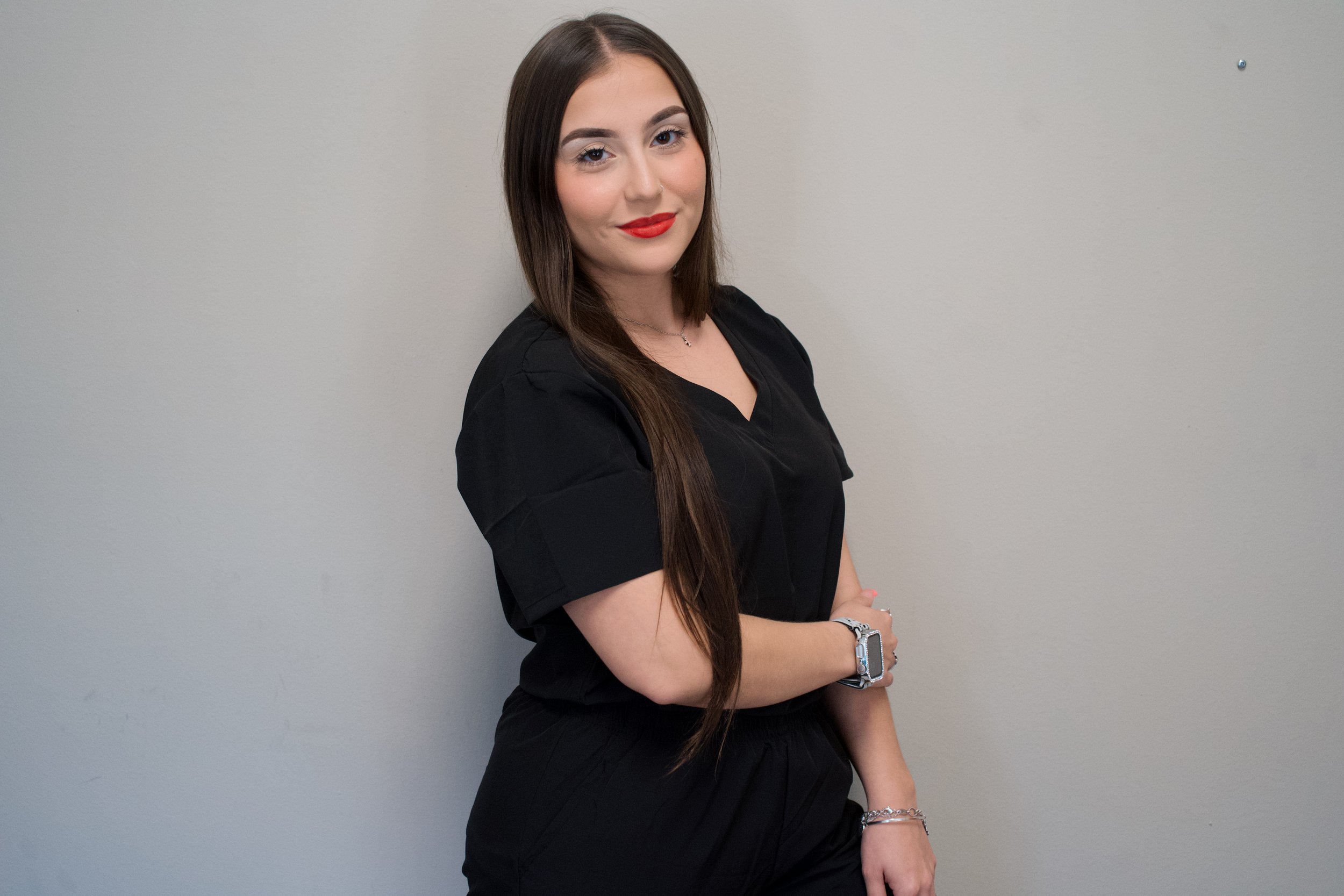 A young woman with long brown hair, wearing a black top and black pants, standing against a plain light gray wall with her arms crossed and smiling.