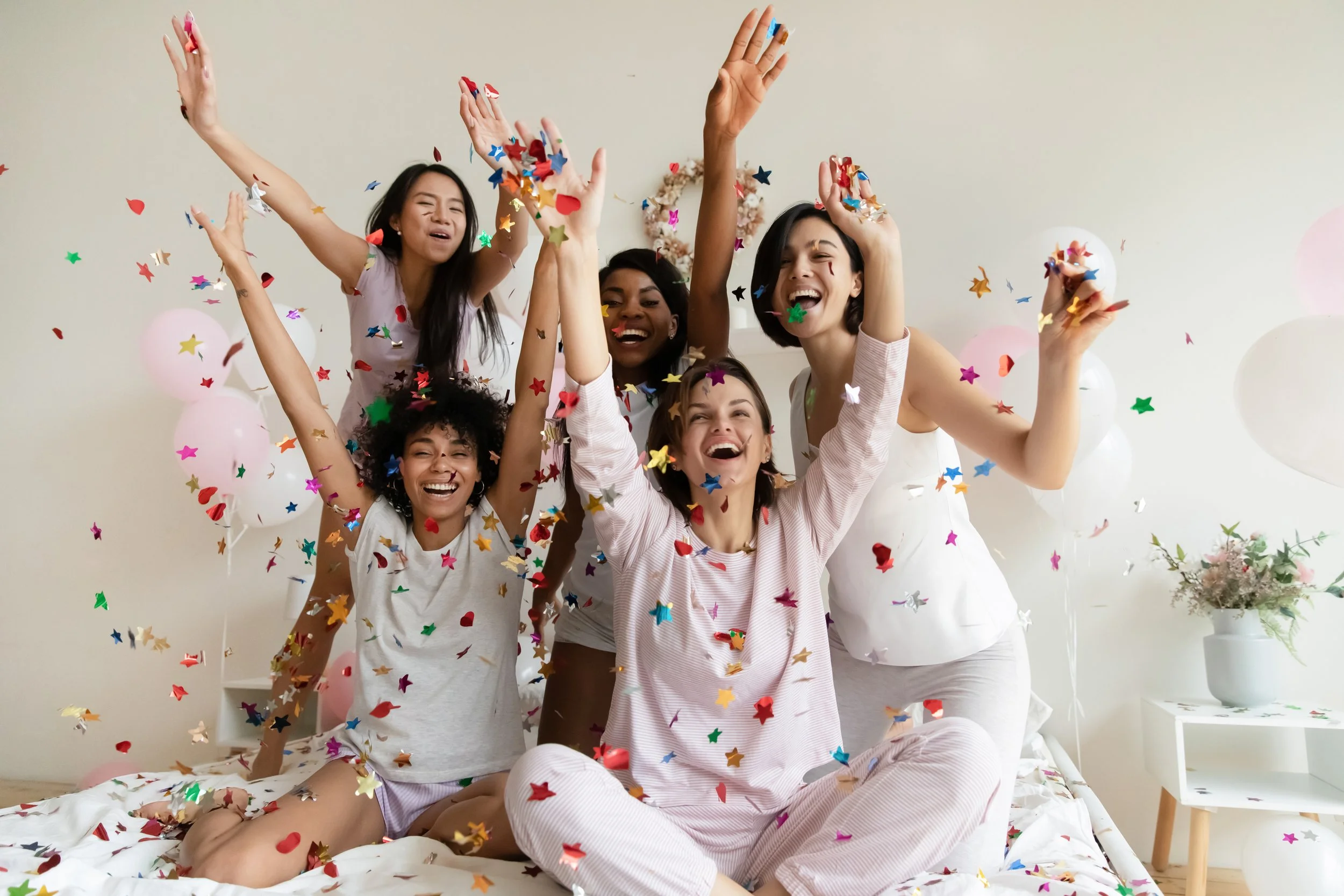 group of five women celebrating with confetti in a bedroom, smiling and raising their hands, with balloons and flowers in the background.