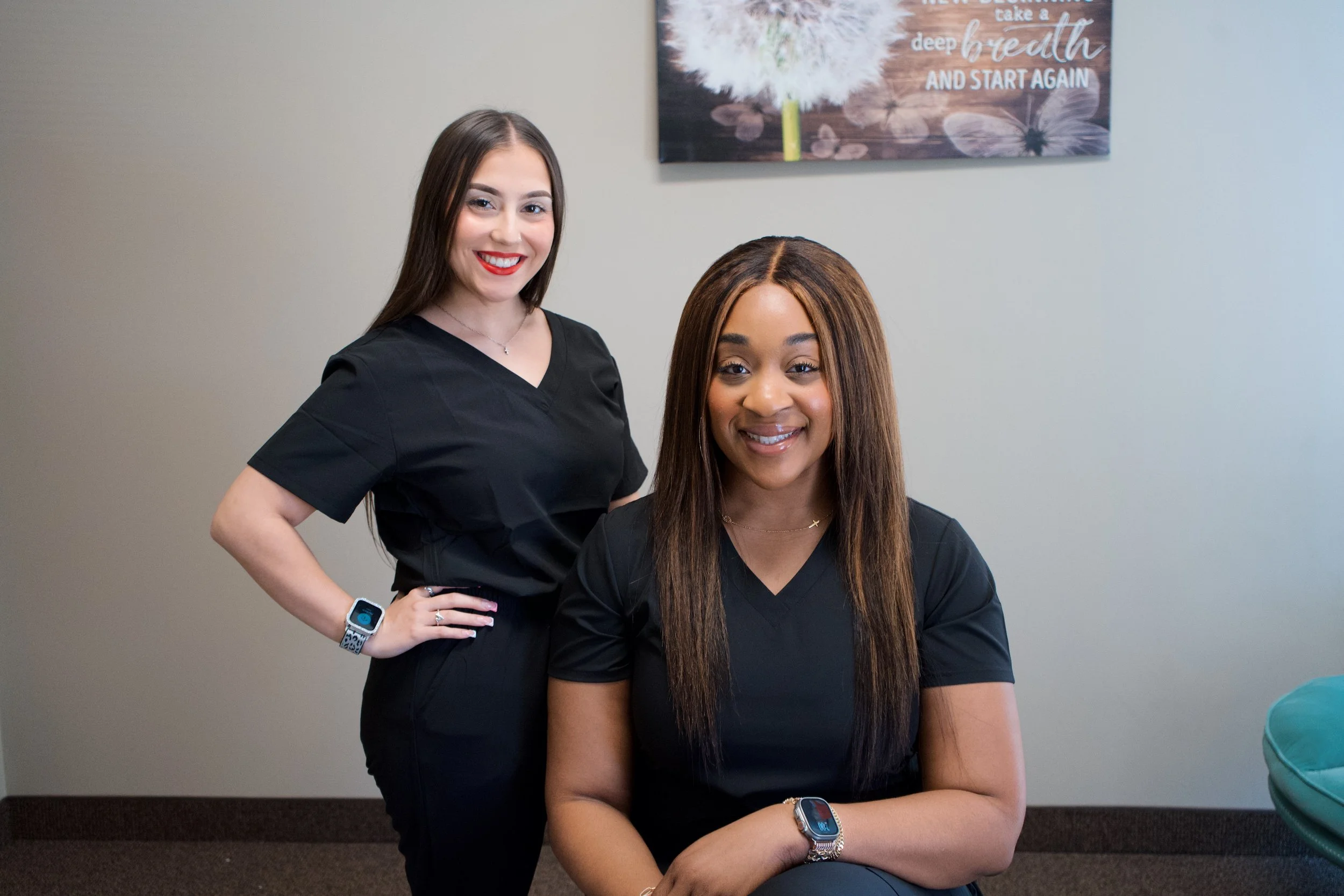 Two women wearing black scrubs smiling indoors, one standing and one seated, with a framed motivational quote on the wall behind them.