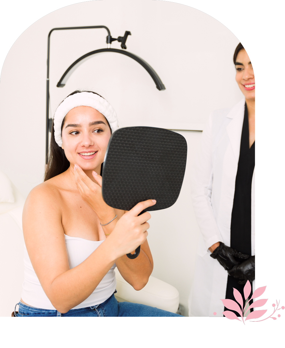 A young woman with a white towel on her head smiling while looking at her reflection in a mirror, with a healthcare professional standing nearby, wearing gloves and a white coat.