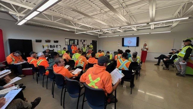 Safety training session with workers in orange and yellow safety vests, seated in a classroom, listening to a presenter near a screen.