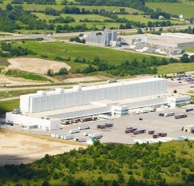 Large industrial building with trucks parked outside, surrounded by green fields and trees.