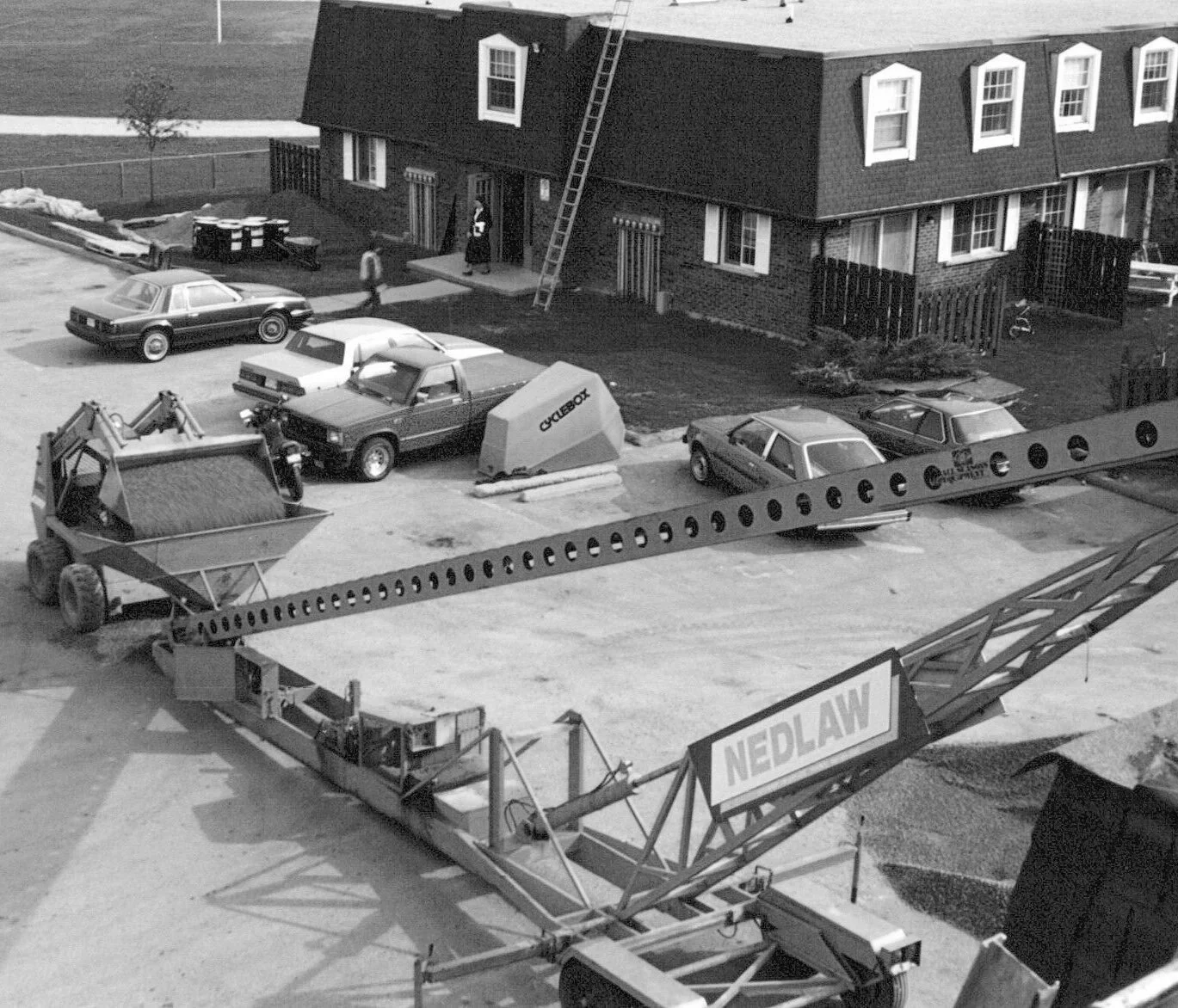 Black and white photograph showing a construction site with a crane labeled NEDLAW, several parked cars, a house with a ladder on the roof, and construction equipment.