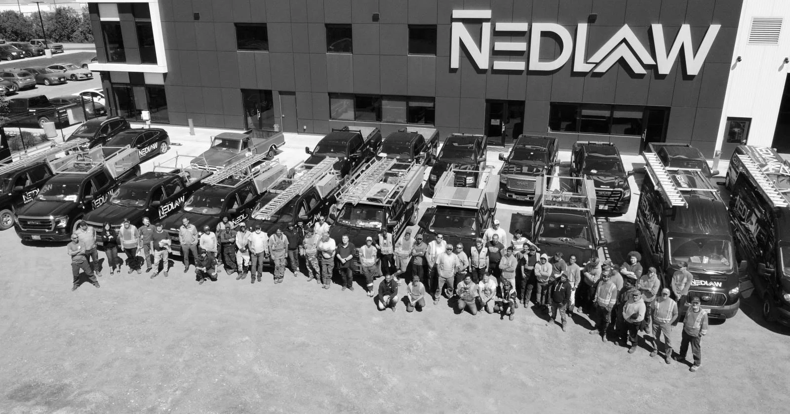 A group of people standing in front of multiple service trucks and cars parked outside a building with a large sign that reads 'NEDLAW'.