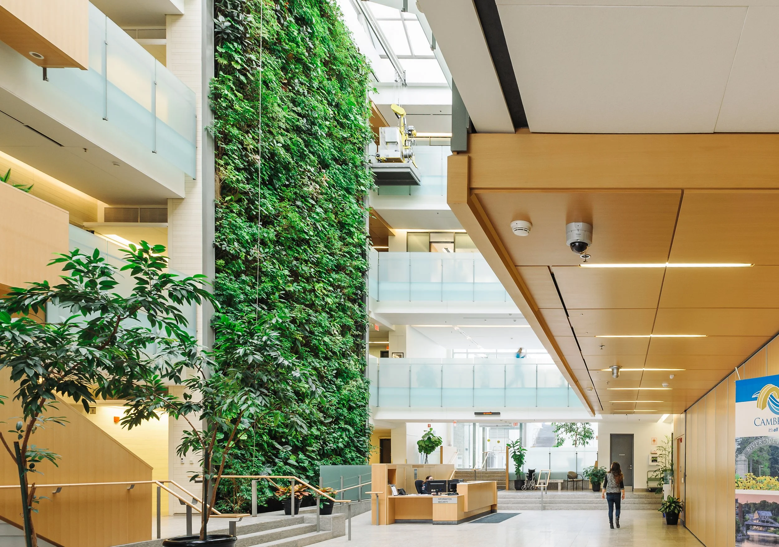 Interior view of a modern building with a large green wall on the left side featuring lush plants, multiple levels with balconies, and a reception area with a woman walking in the foreground.