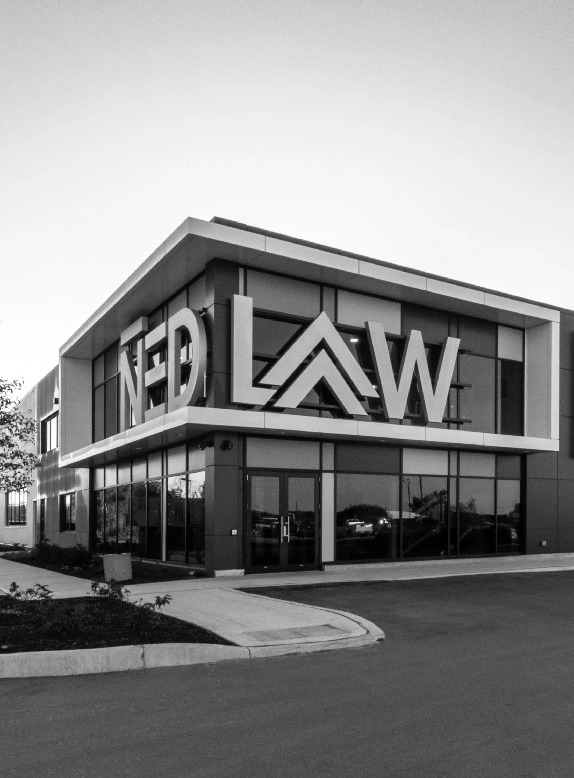 Black-and-white photo of a modern office building with large windows and a sign on the corner that reads 'NED LAW'. The building is surrounded by a parking lot and sidewalk.