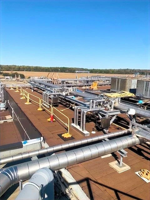 Industrial rooftop with various pipes, valves, and safety railings under a clear blue sky.