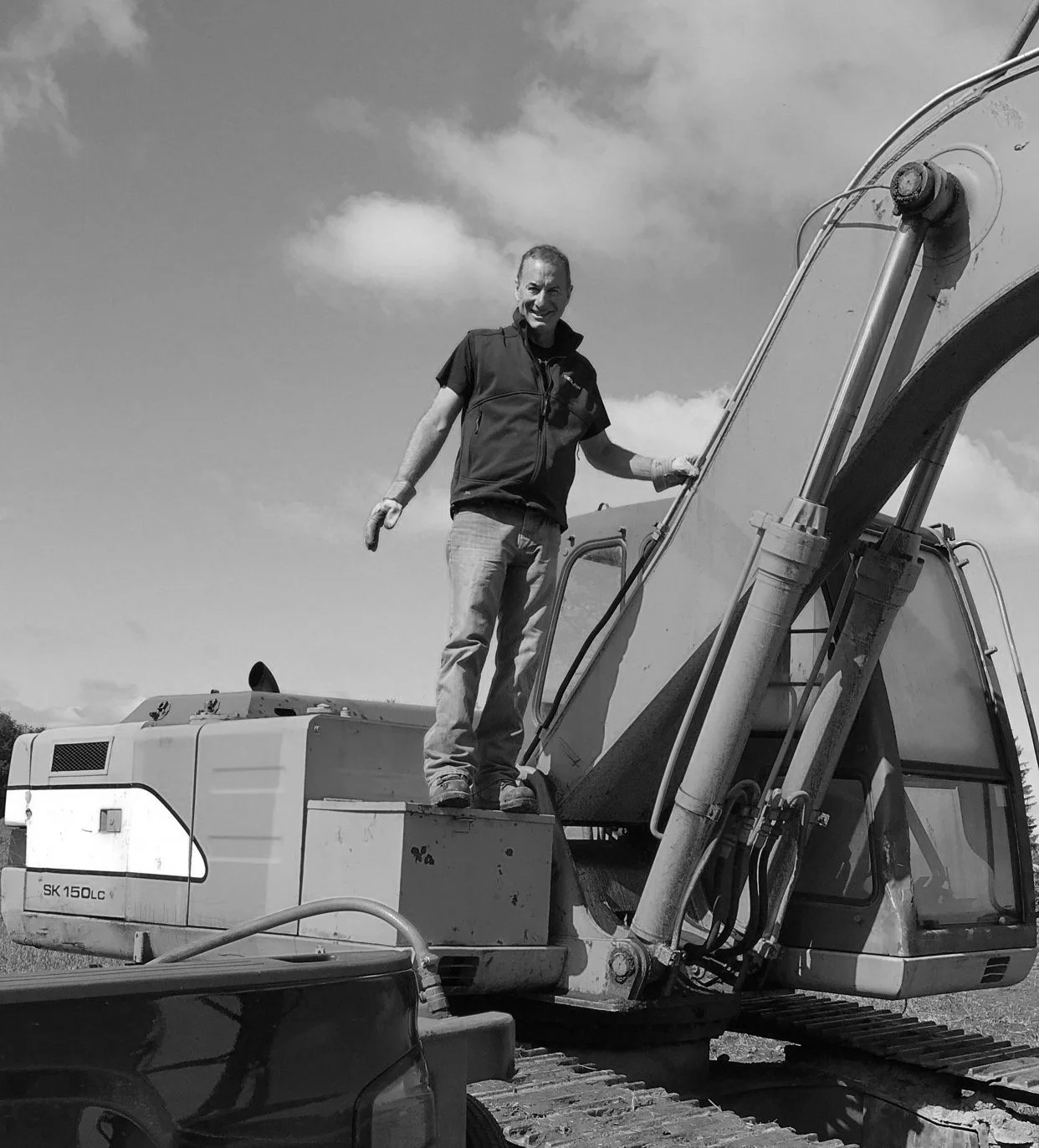 A man standing on an excavator with a smile, holding the operator's handrail, outdoors under a sky with clouds.
