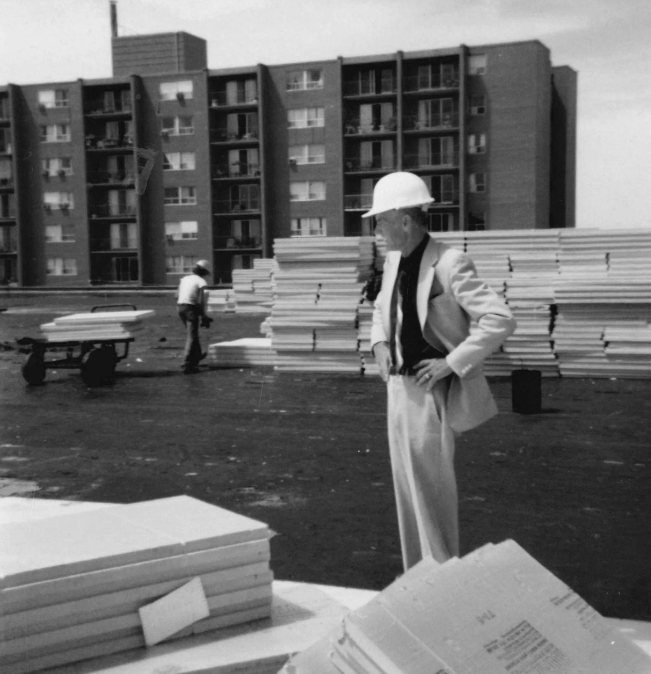 A man wearing a hard hat and light-colored coat sitting on a bench near stacks of white bricks in an outdoor urban setting with a tall apartment building in the background.