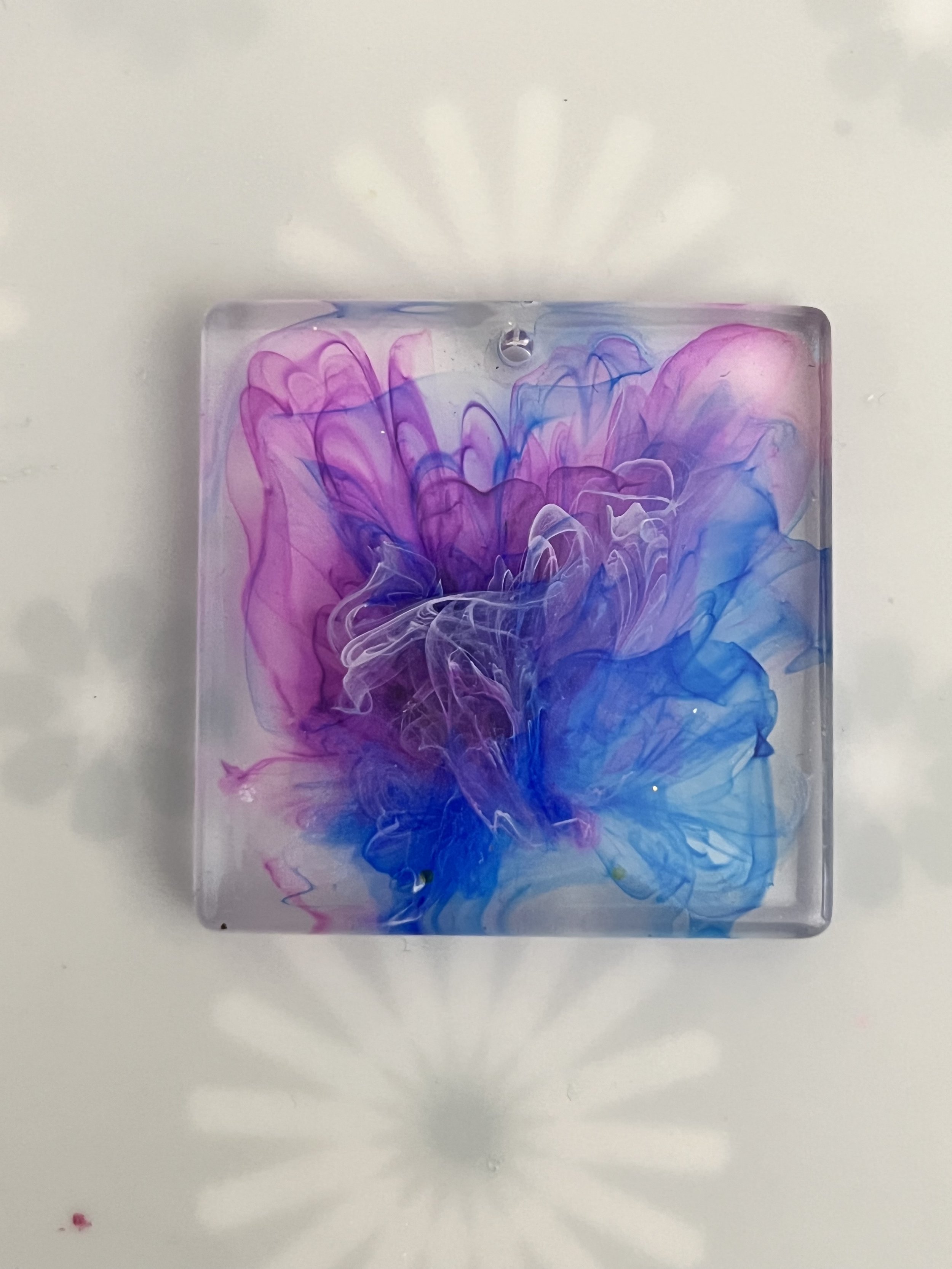 Colorful ink swirling in water inside a transparent square container, creating a vibrant abstract pattern with shades of purple, pink, and blue, casting a shadow on a white surface.