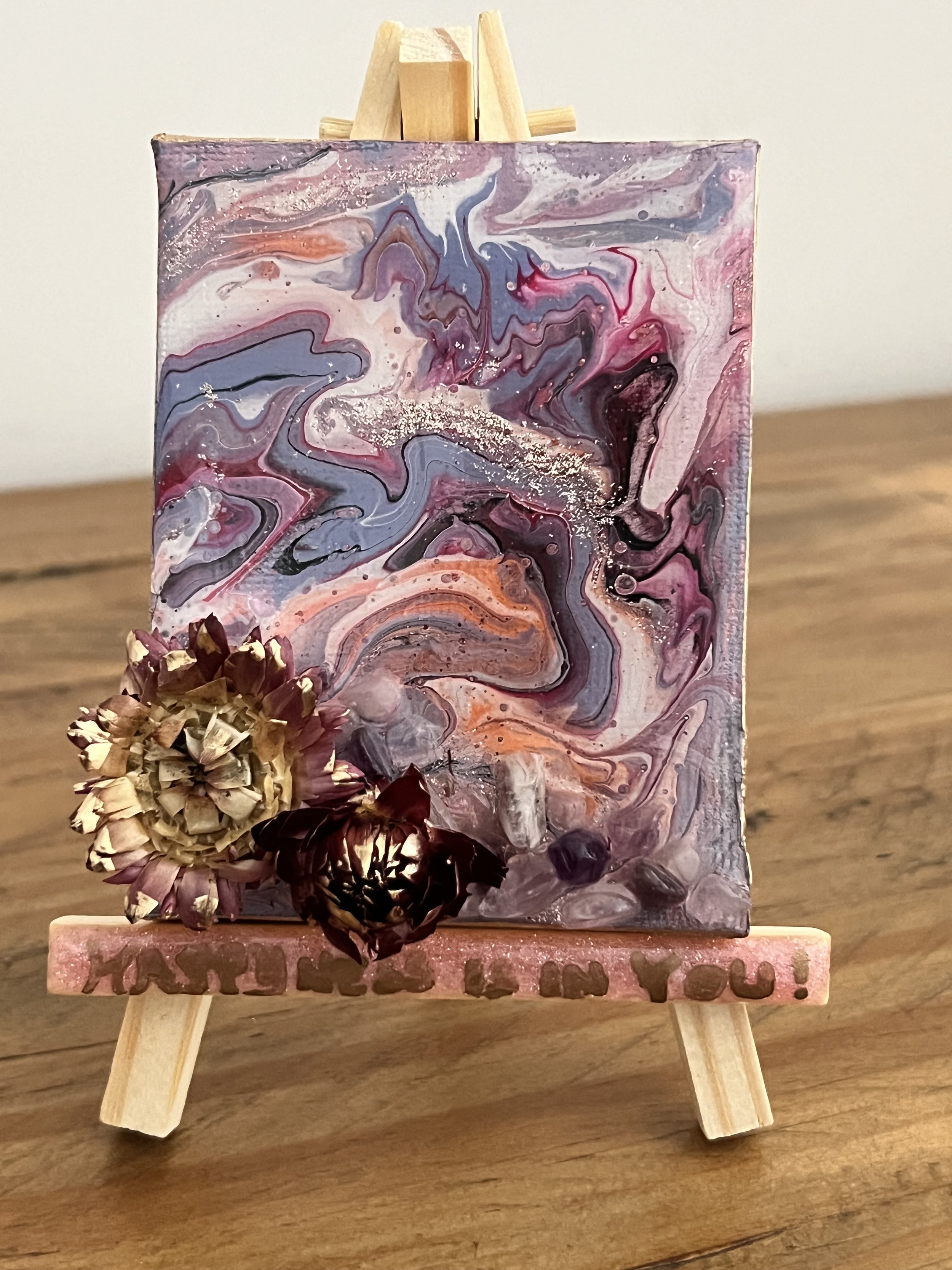 Abstract marbled painting with swirls of purple, pink, orange, and black, adorned with three dried flowers at the bottom left, placed on a small pink easel with the phrase "Mother's Day in You!" engraved at the base.