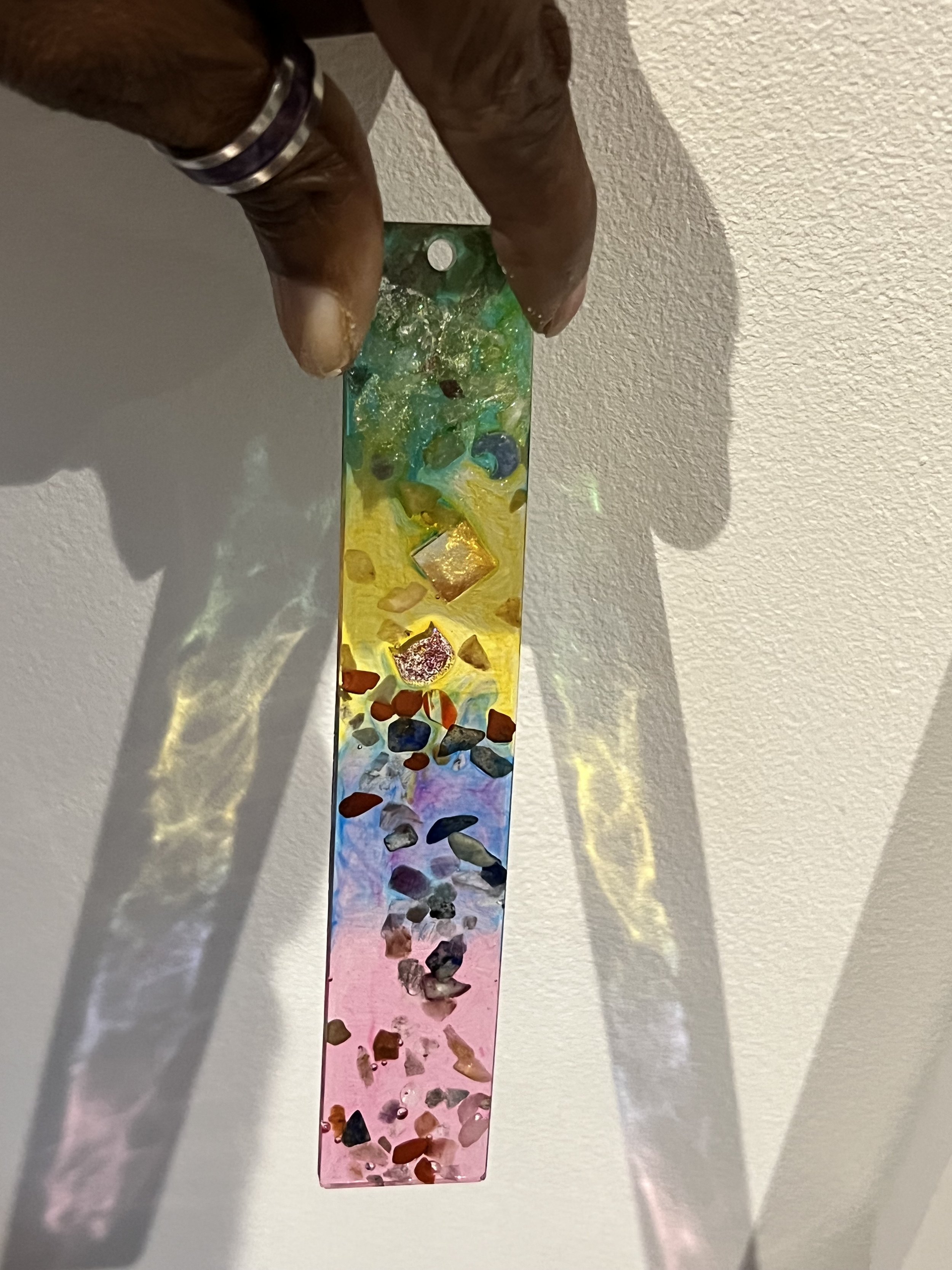 A person holding a colorful, rectangular art piece made of glass with embedded small stones, casting a rainbow shadow on a white wall.