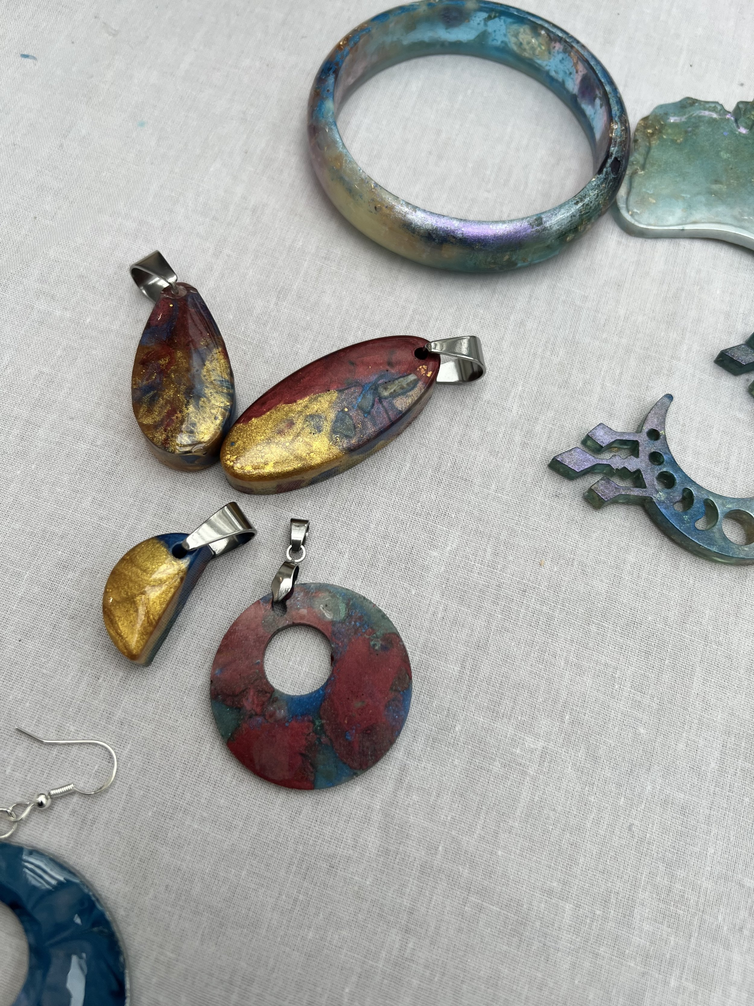 Colorful handmade jewelry including pendants, a bracelet, and a crescent-shaped piece displayed on a white fabric surface.
