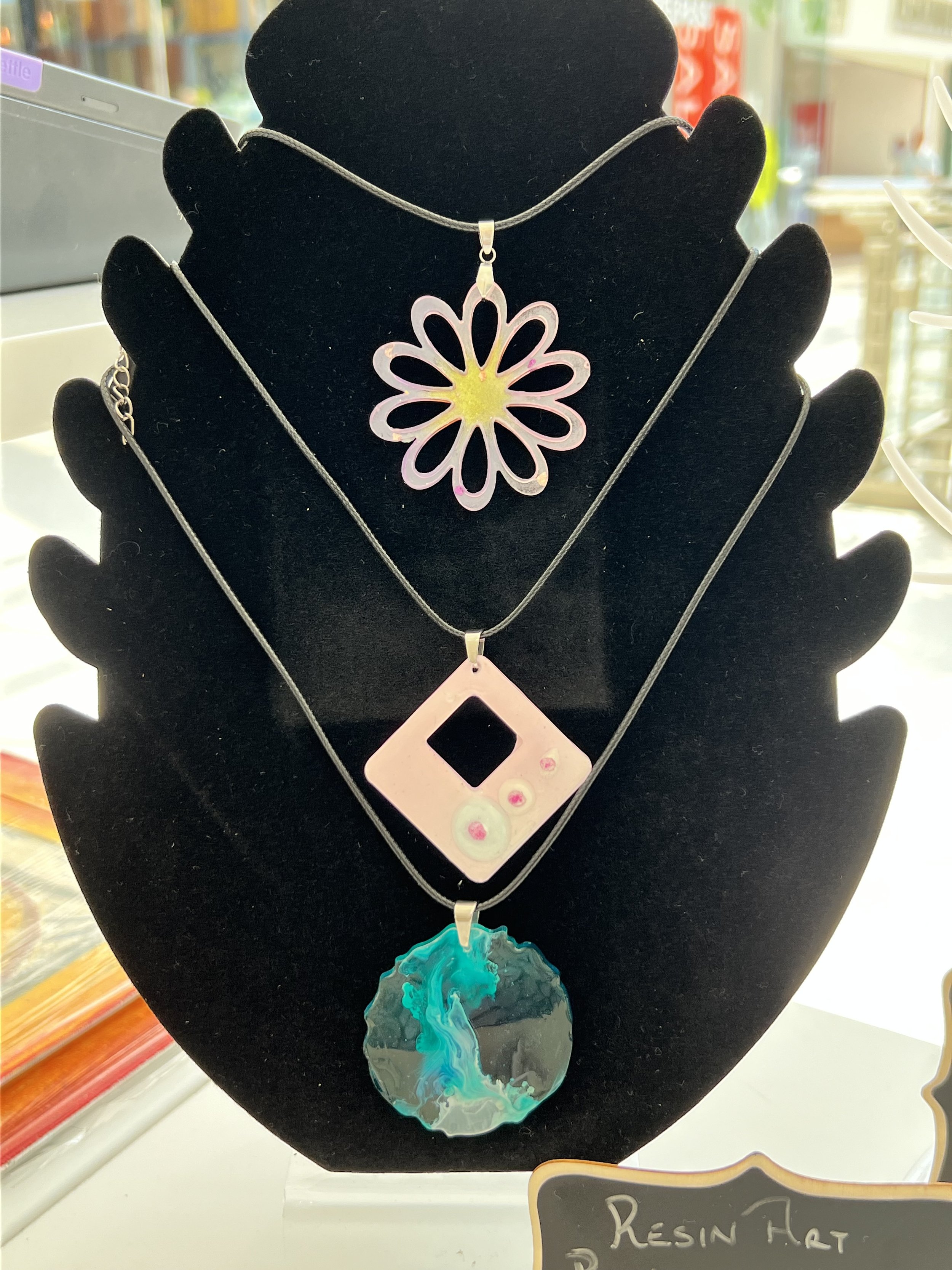 Three necklaces with resin pendants displayed on a black jewelry stand. The top pendant is a flower-shaped design, the middle pendant is a pink square with a cutout and small spheres, and the bottom pendant is a circular disc with abstract blue and g