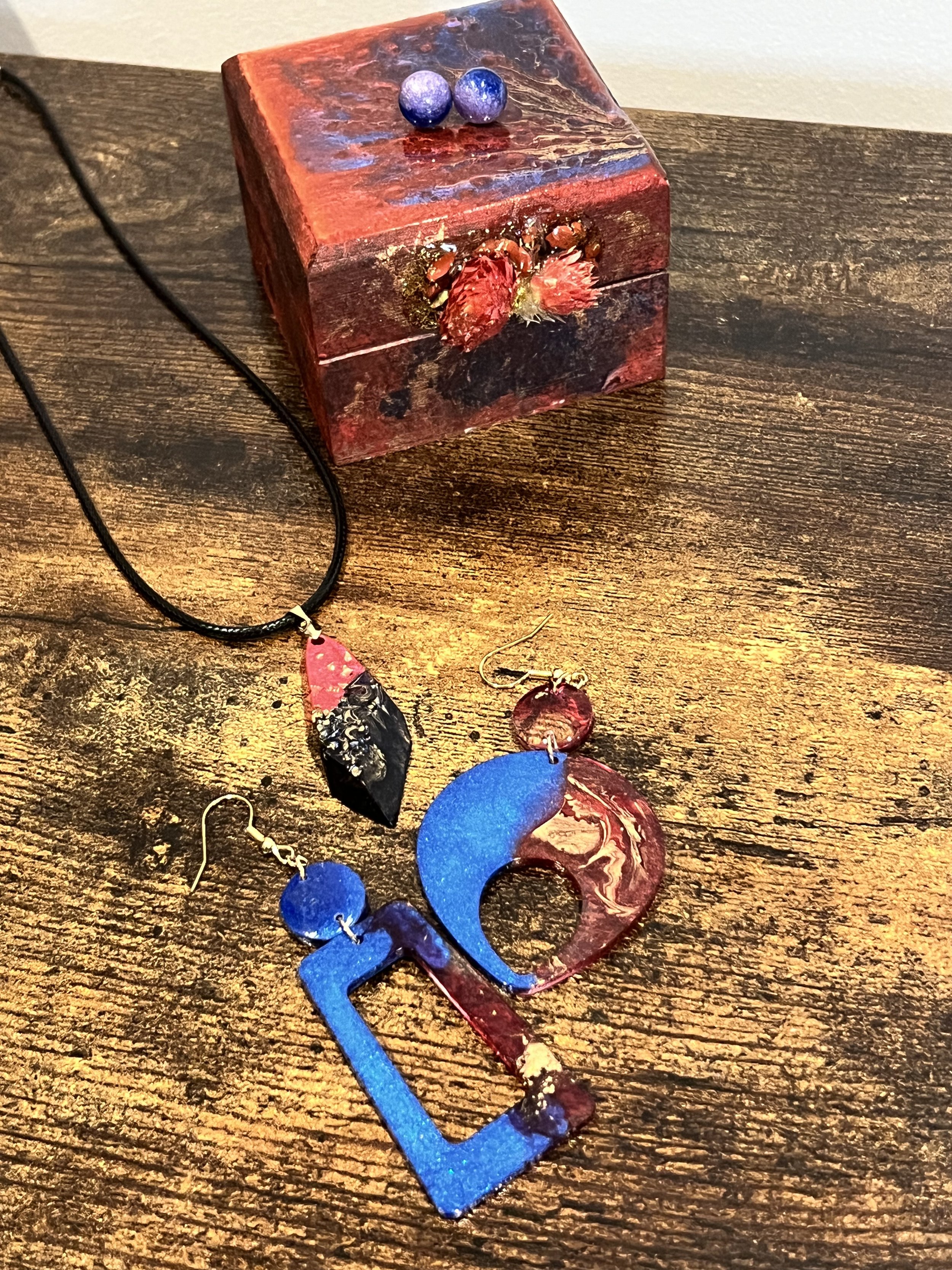 Colorful jewelry set including a necklace and earrings, with a wooden box in the background. The necklace has a black cord and a pendant with red and black sections, and the earrings are designed with blue, red, and multicolored elements. The wooden