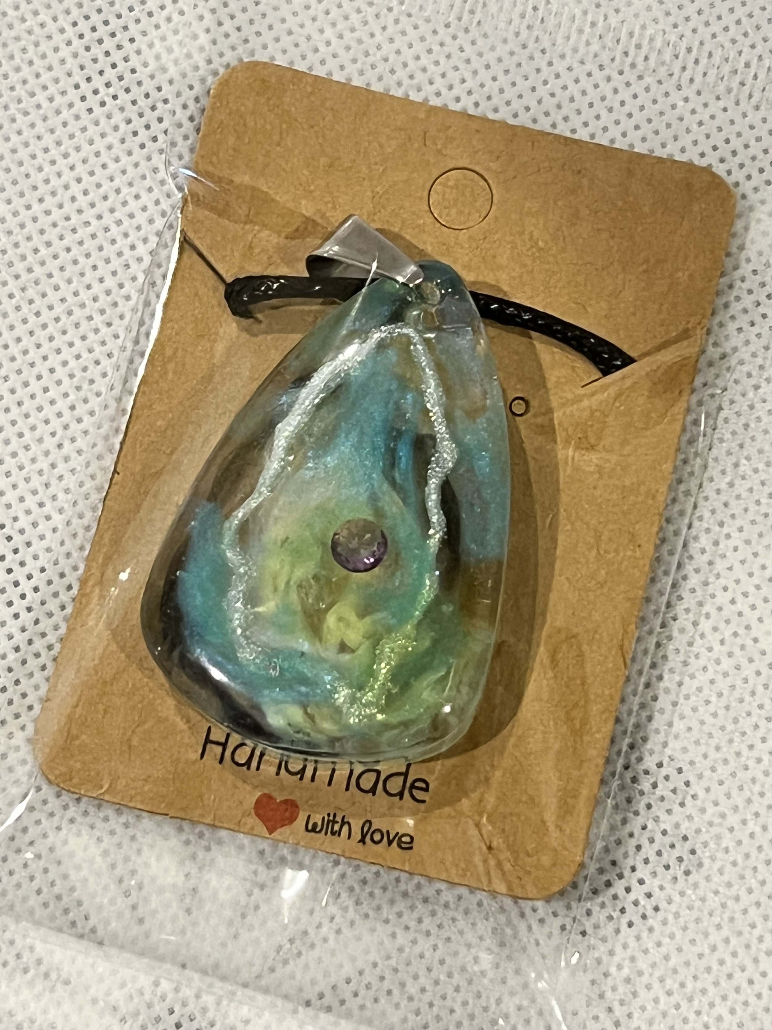 Colorful glass pendant in shades of blue, green, and yellow with a purple accent, attached to a black cord, on a brown paper card that reads "Handmade with love."