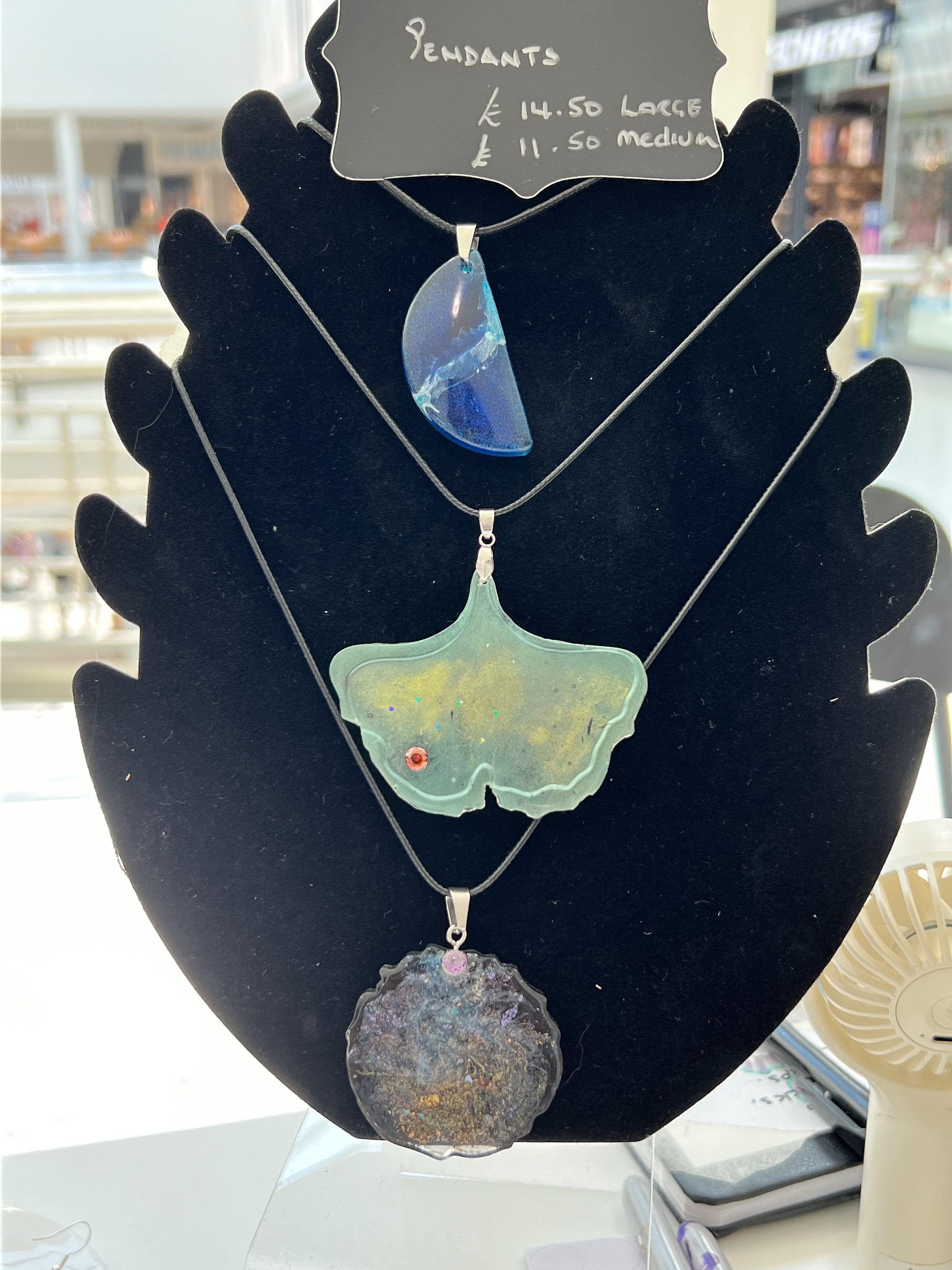 Display of three glass pendant necklaces on a black jewelry stand, with pricing signs indicating large and medium sizes.