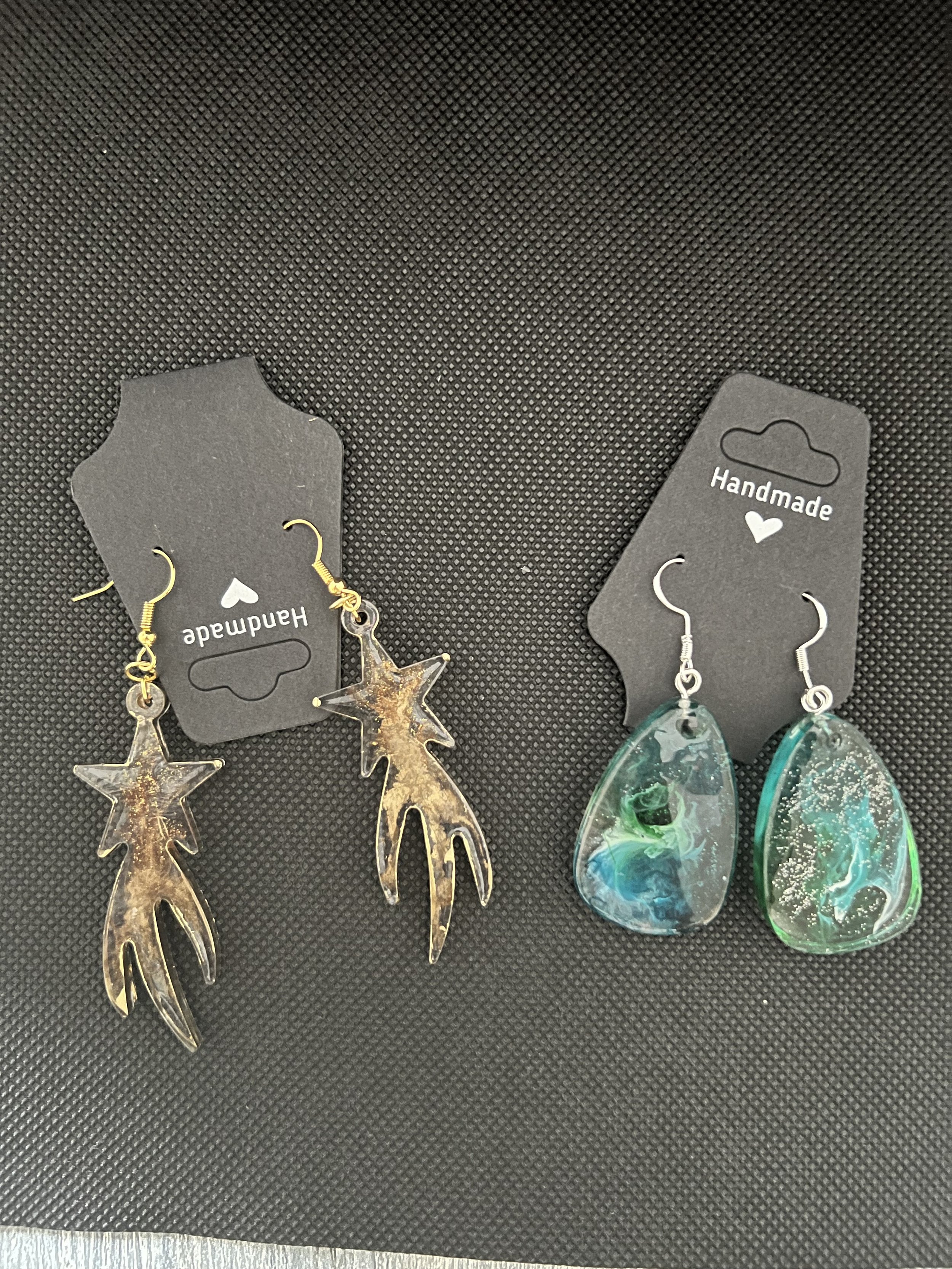 Three pairs of handmade earrings on black display cards, featuring star-shaped, teardrop-shaped, and abstract colorful designs.
