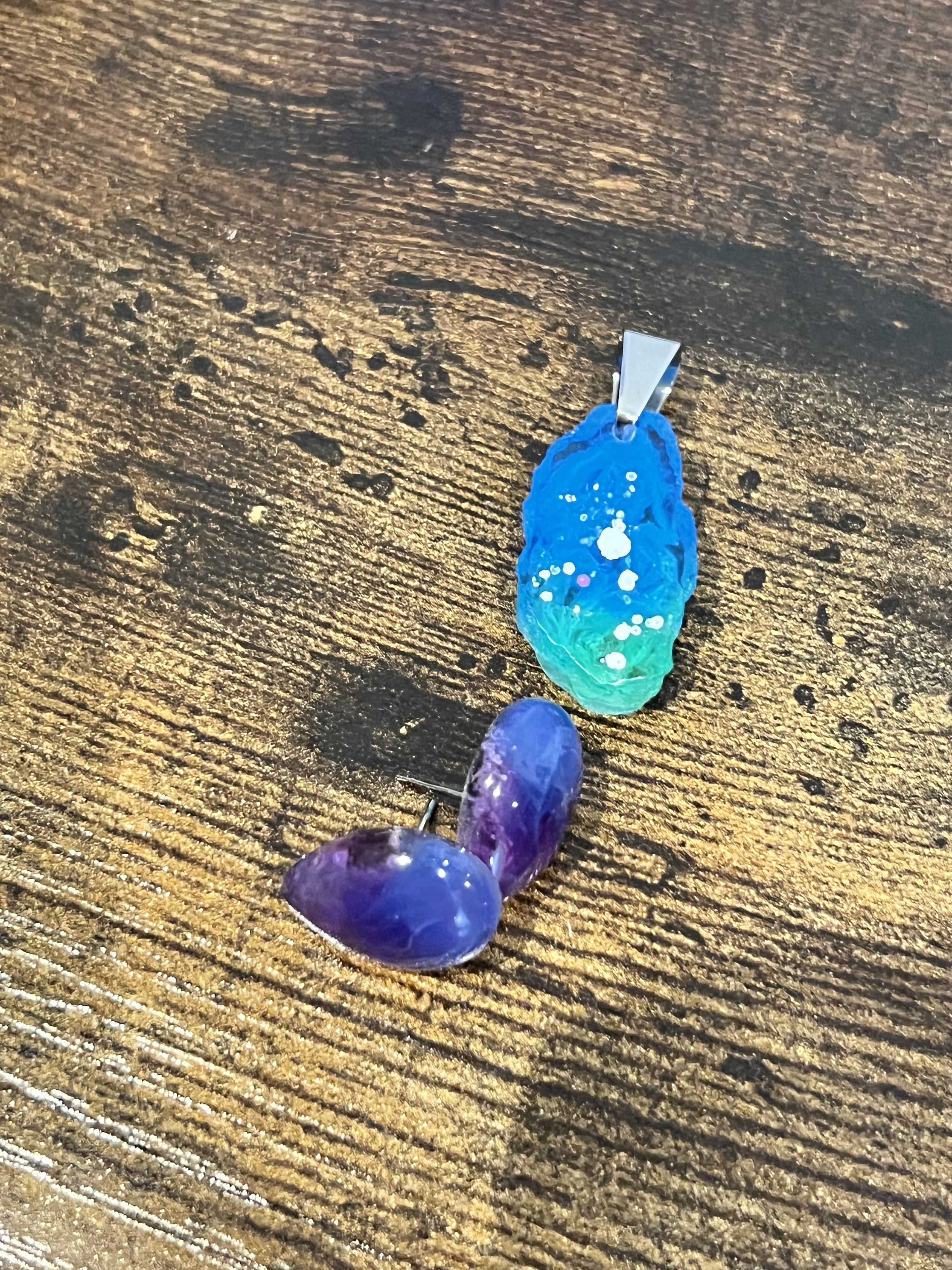 Two colorful resin pendants, one blue with white spots and one purple with a glossy finish, resting on a wooden surface.