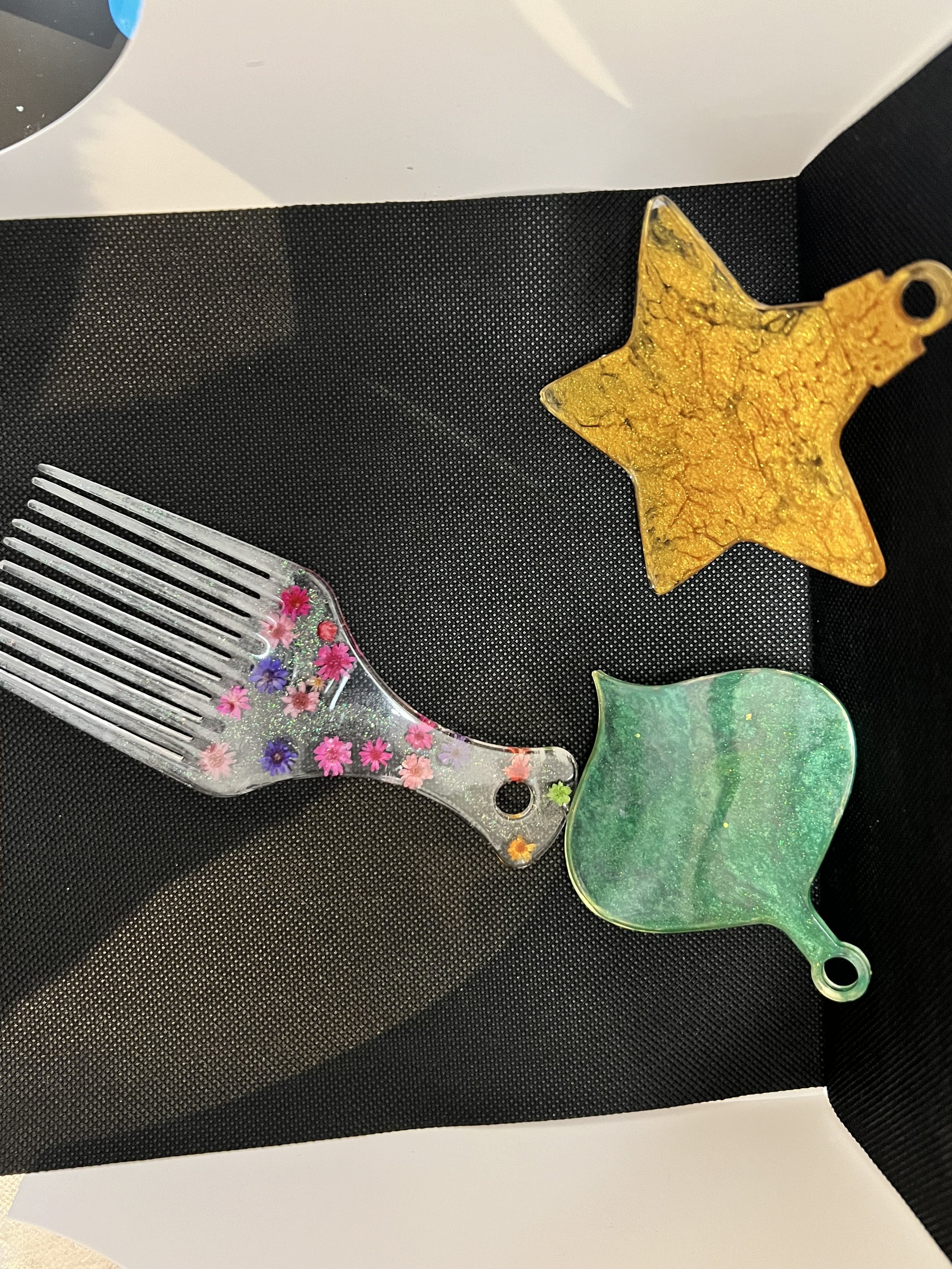 Three decorative glass ornaments in shapes of a star, a leaf, and a fan, with colorful patterns and glitter, resting on a black textured surface.
