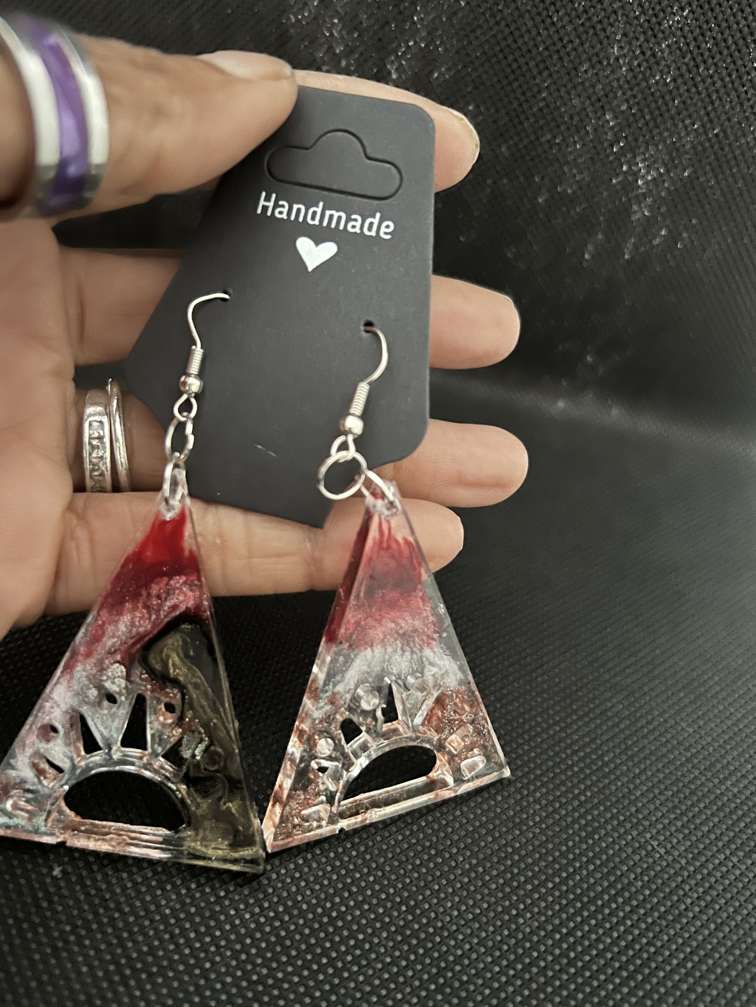 A pair of handmade triangular earrings with a decorative cutout and red, black, and white marble-like design, attached to a black display card labeled 'Handmade' with a small heart symbol, being held by a person with rings on their fingers.
