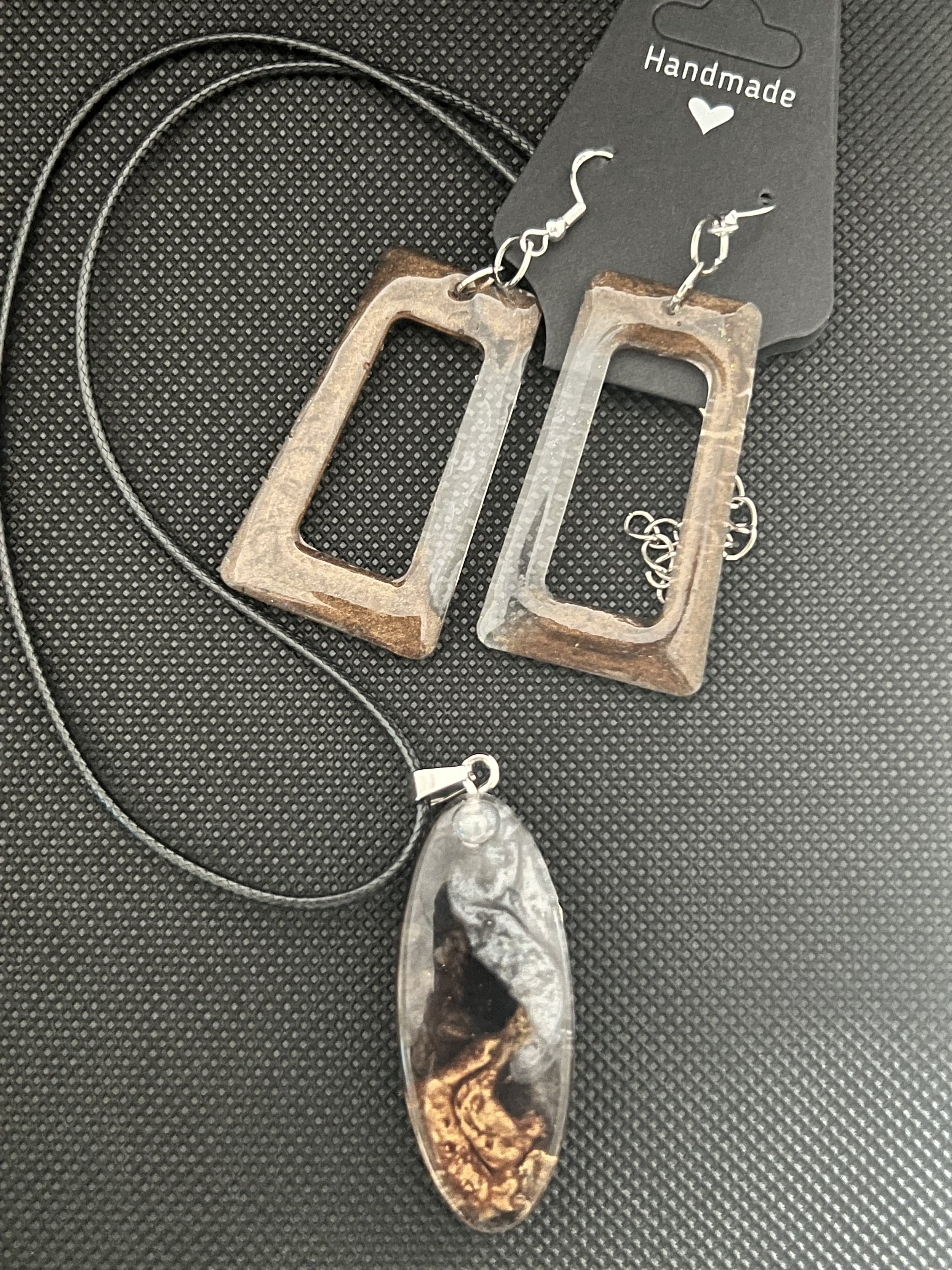 A pair of handmade earrings with rectangular, open frame design, a black and gold oval pendant necklace on a black textured surface.